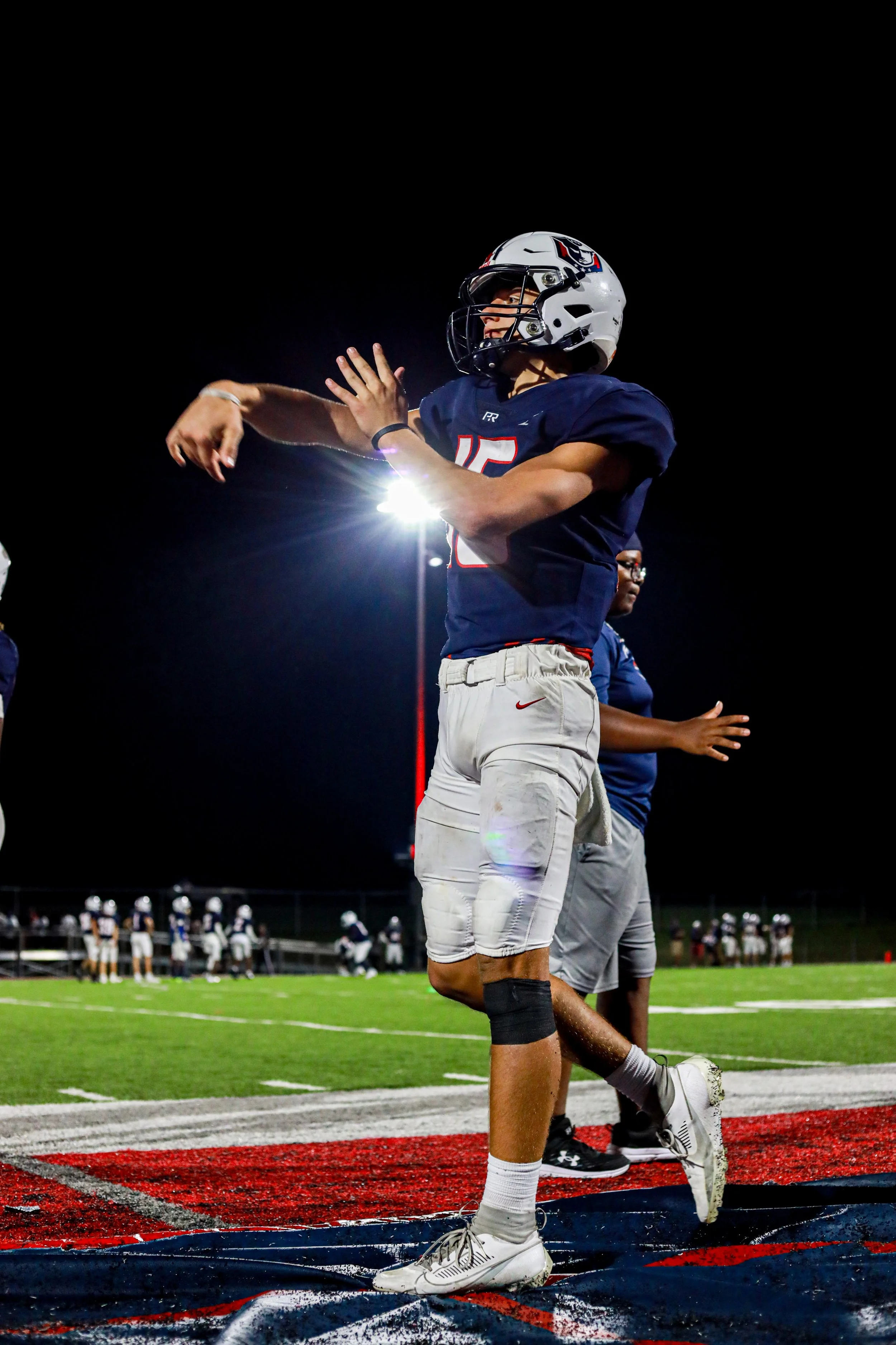 High school football quarterback in a navy jersey and white helmet kneeling in the end zone under stadium lights