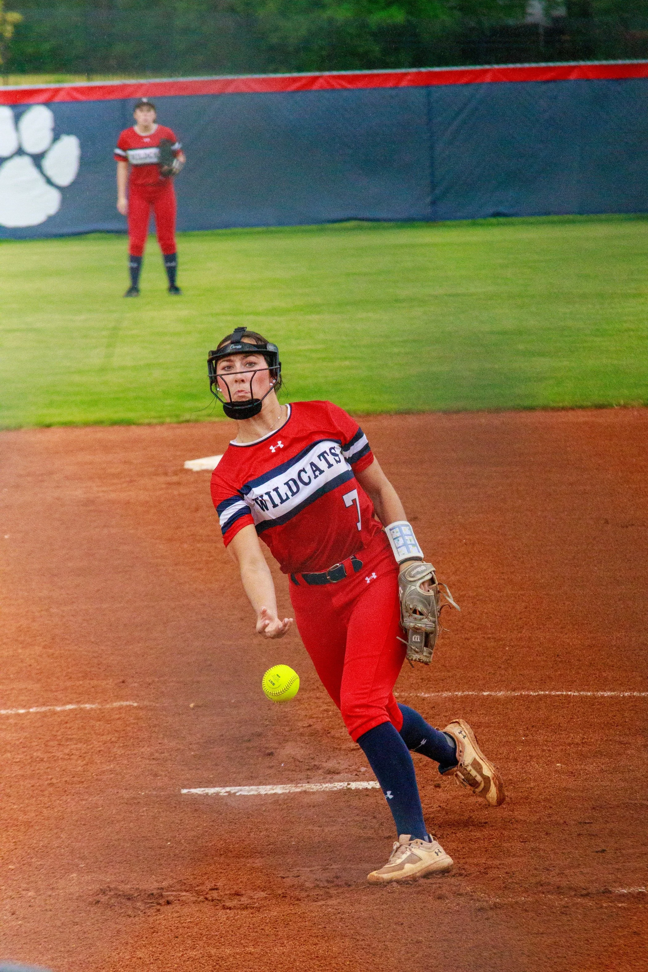 Softball pitcher in a red Wildcats uniform throwing a yellow softball on the mound