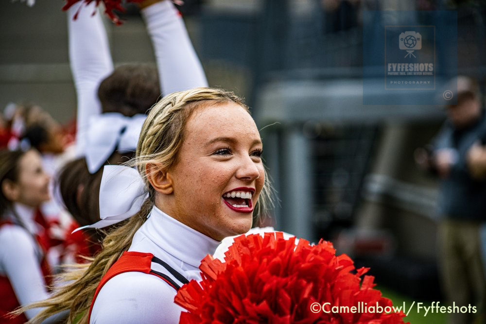 Smiling blonde cheerleader holding red pom-poms during a football game