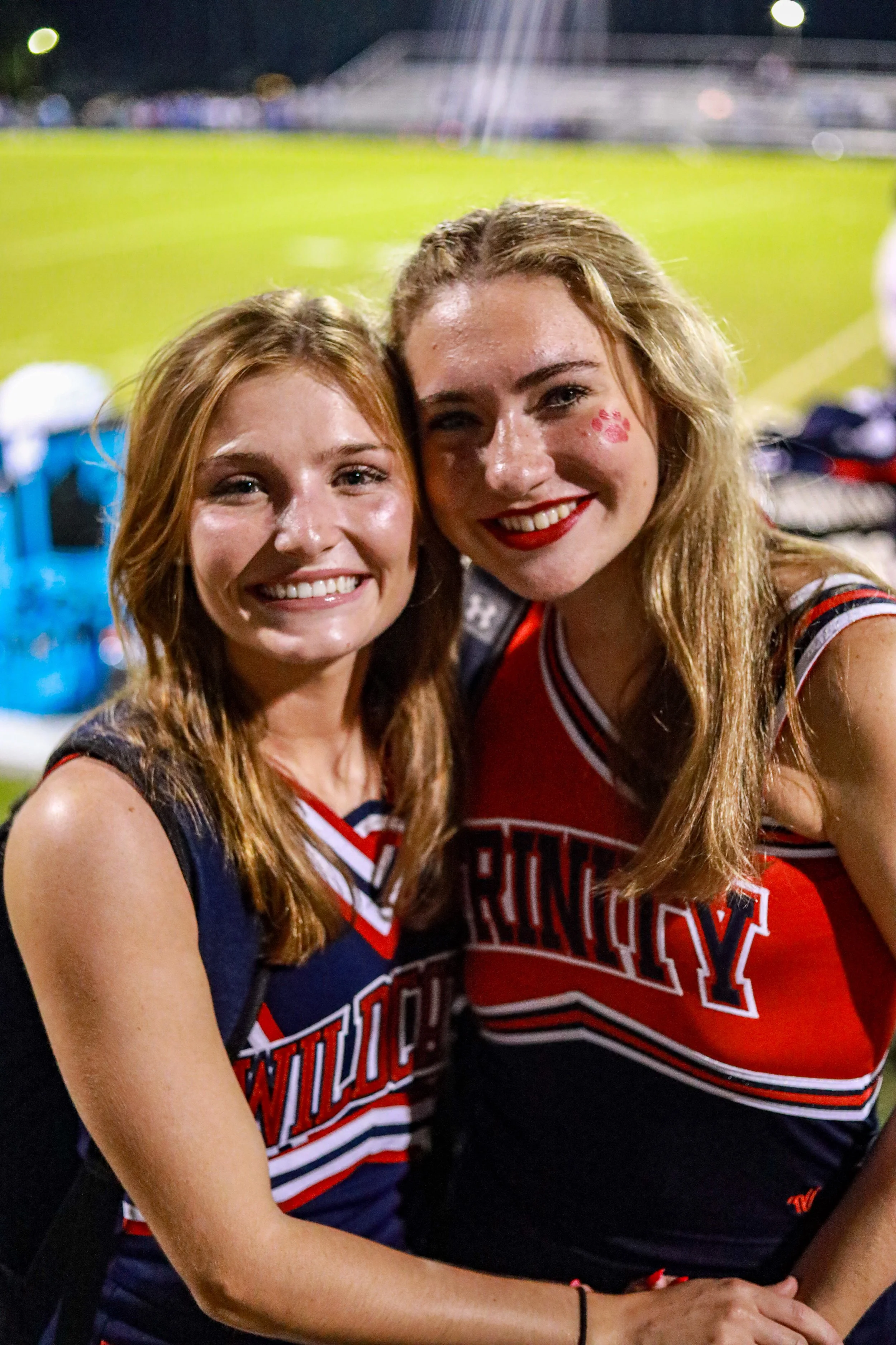 Two smiling women in red, white, and navy cheerleading uniforms pose together at a nighttime event.