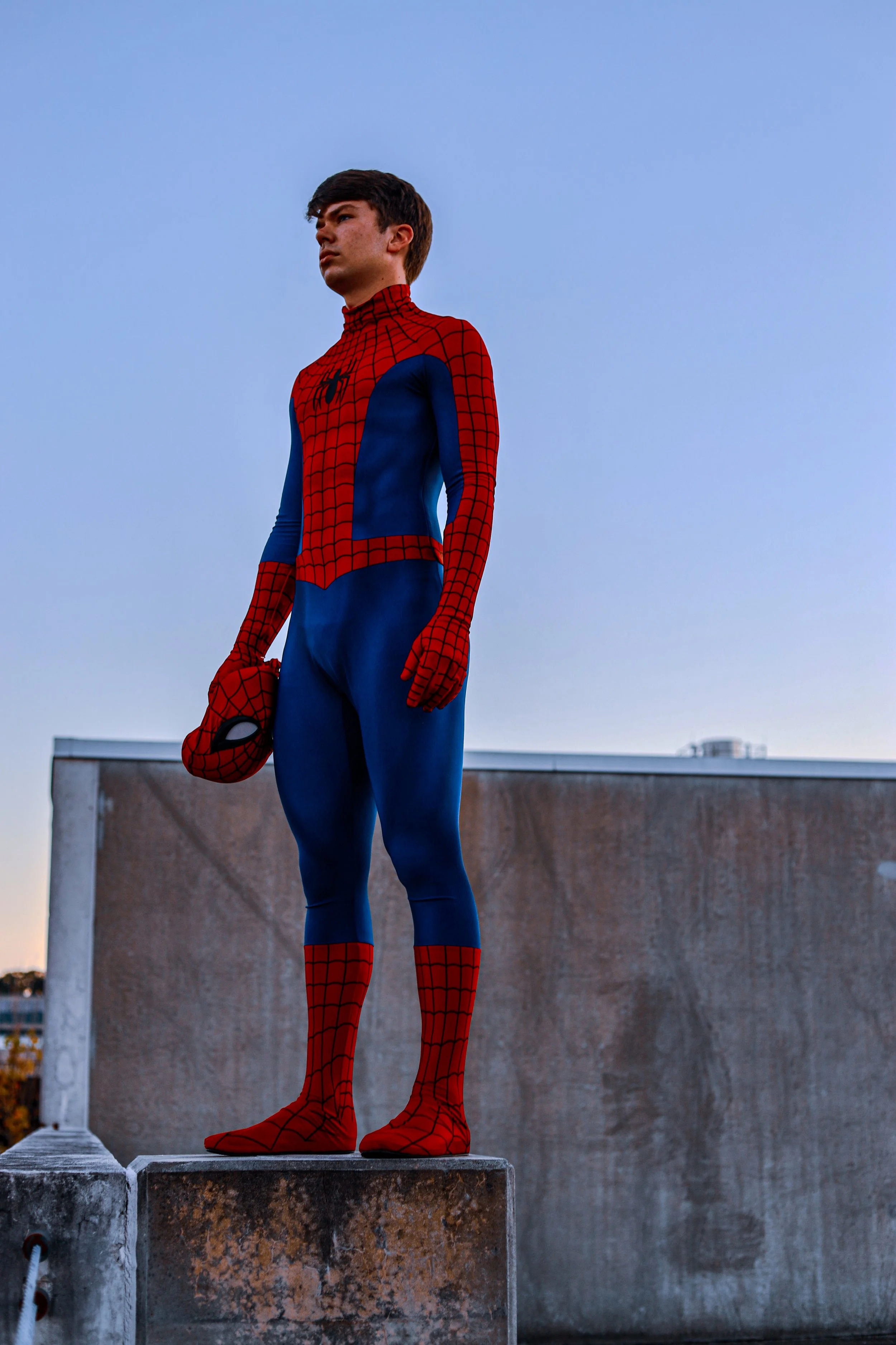Person wearing a red-and-blue Spider-Man costume and holding a Spider-Man mask on a rooftop.