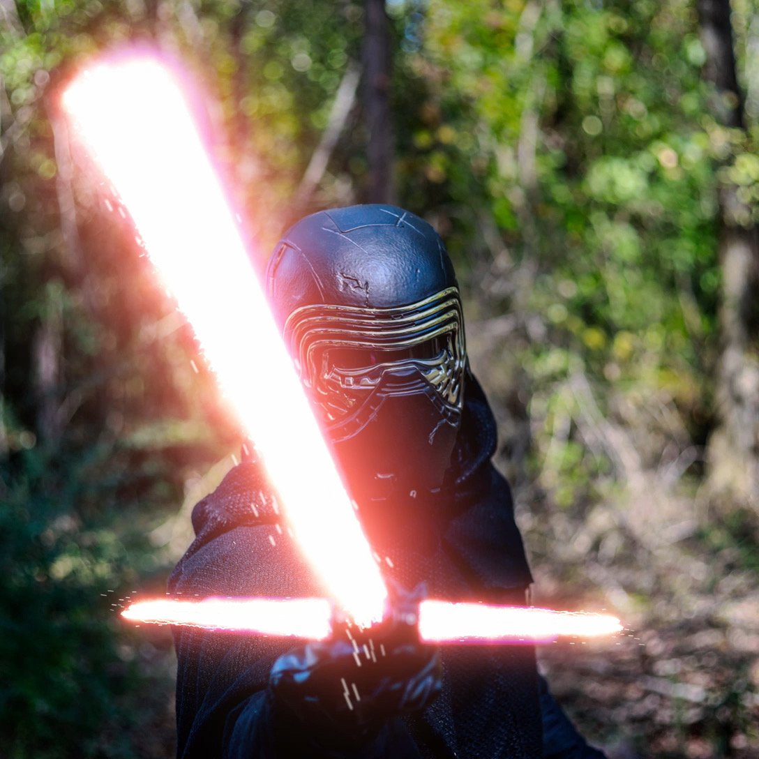 Person in a Kylo Ren costume wielding a glowing red crossguard lightsaber in a forest