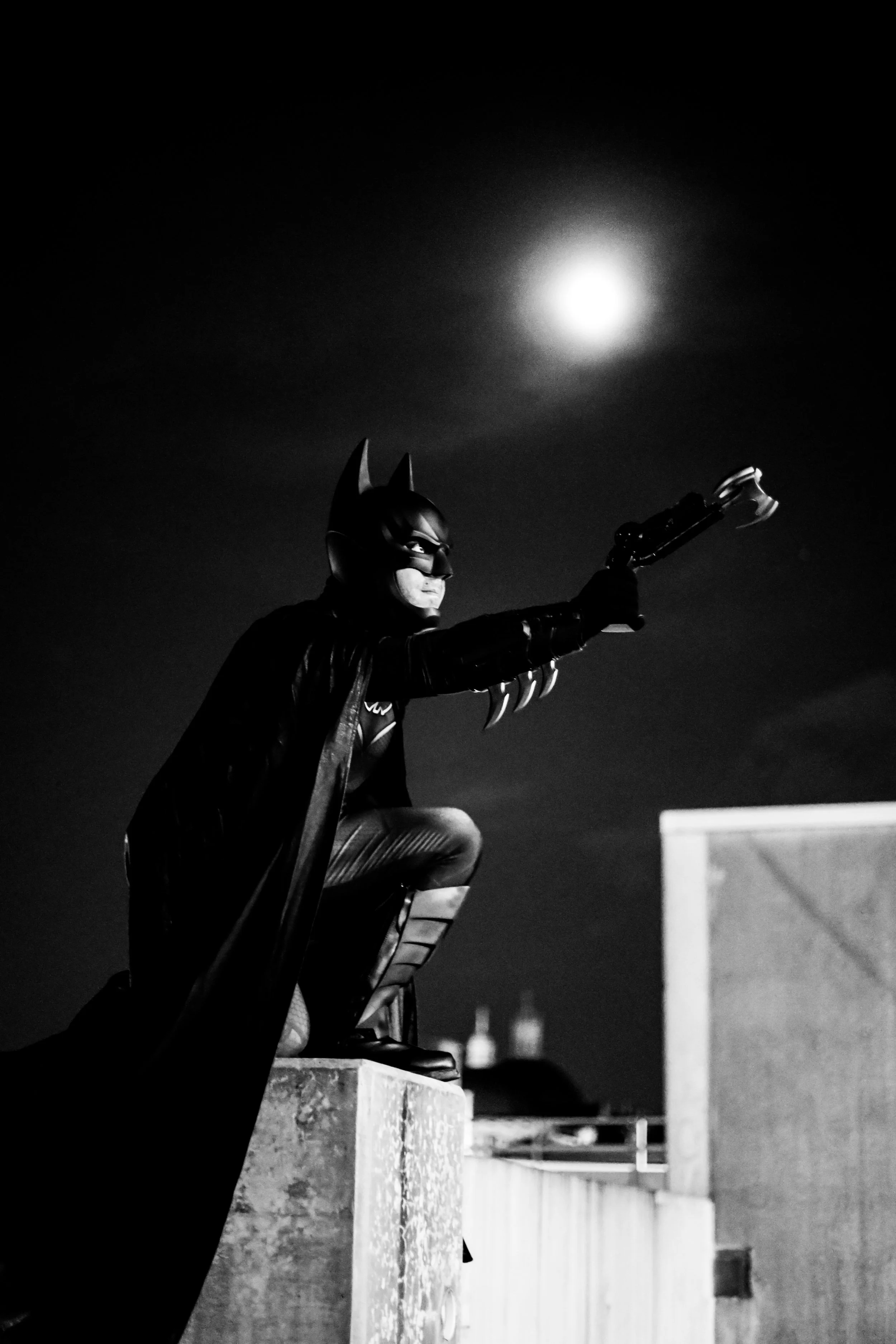 Black-and-white image of Batman kneeling on a rooftop and holding a grappling gun beneath a bright moon.