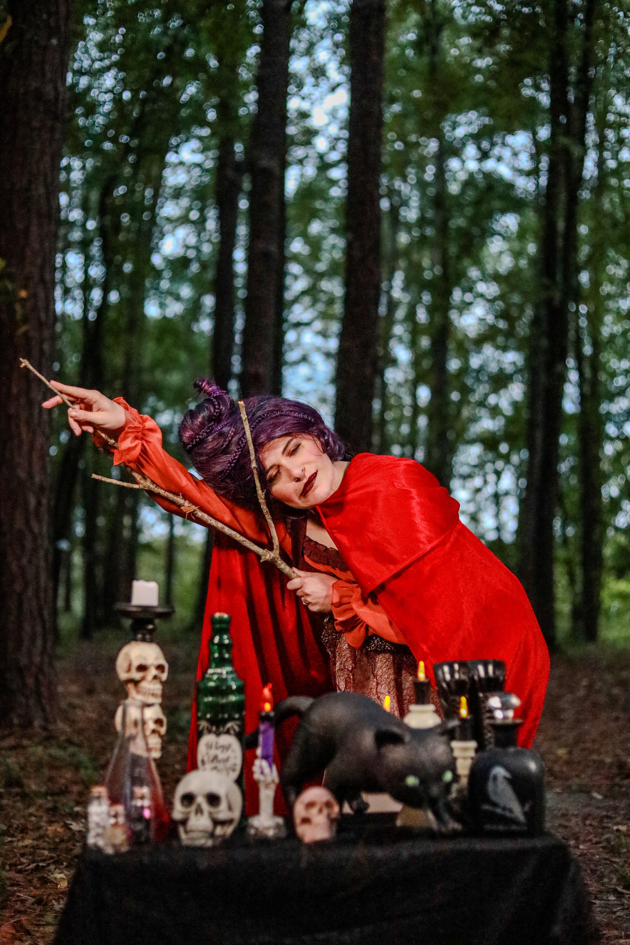Woman in a red cloak holding a branch beside a Halloween altar decorated with skulls, candles, and bottles in a forest.