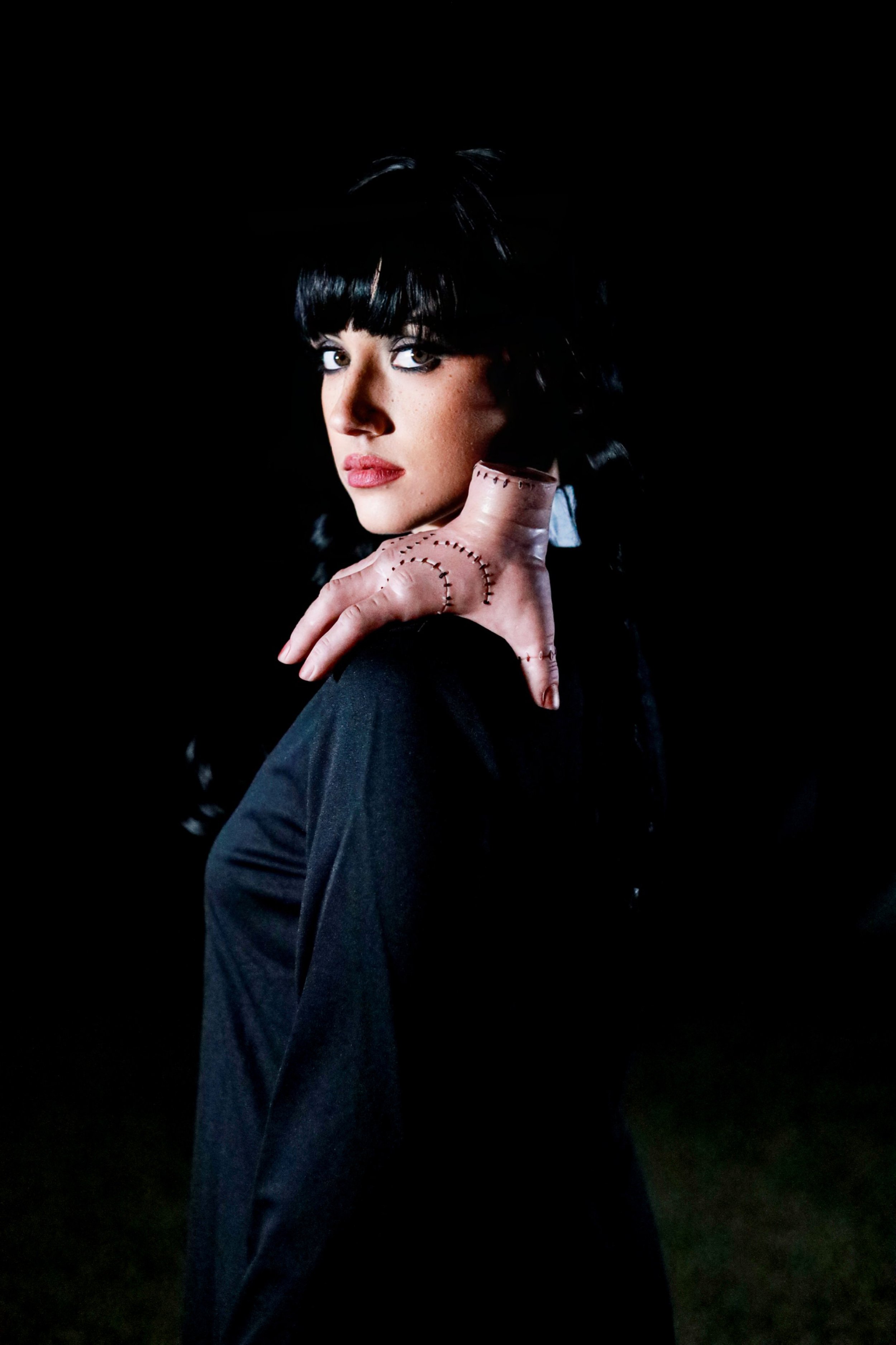 Dramatic portrait of a woman with black hair and bangs, heavy eyeliner, and a stitched pink prosthetic hand against a black background