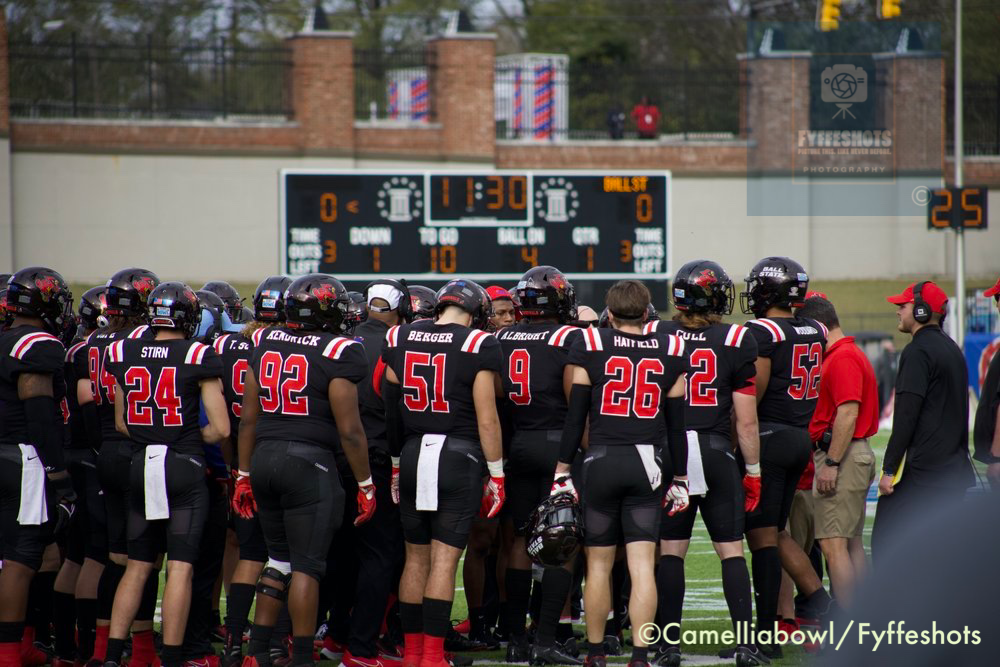 College football team in black and red uniforms huddles on the sideline beneath a scoreboard