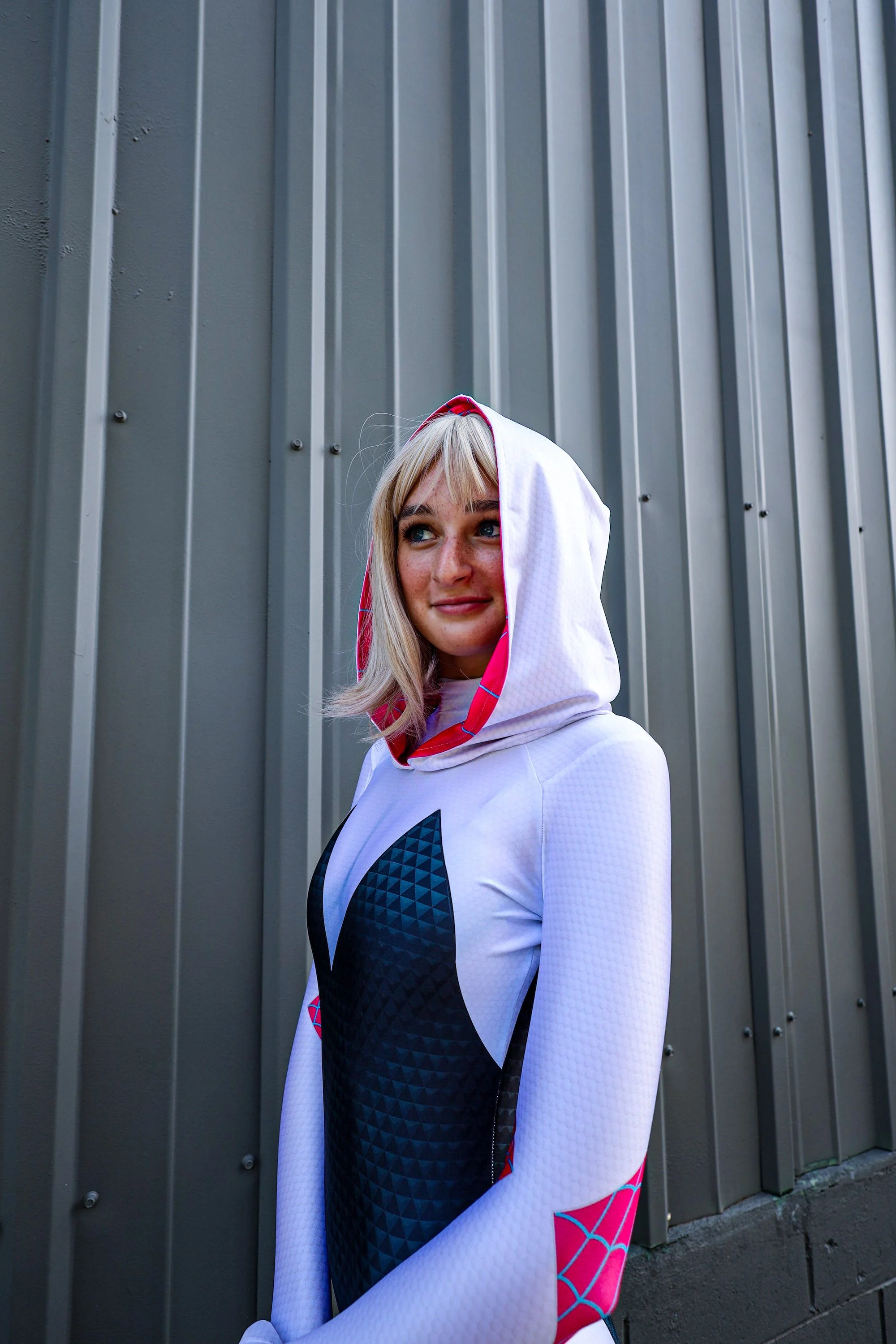 Blonde woman wearing a white and black geometric-patterned hooded outfit standing beside a gray corrugated metal wall