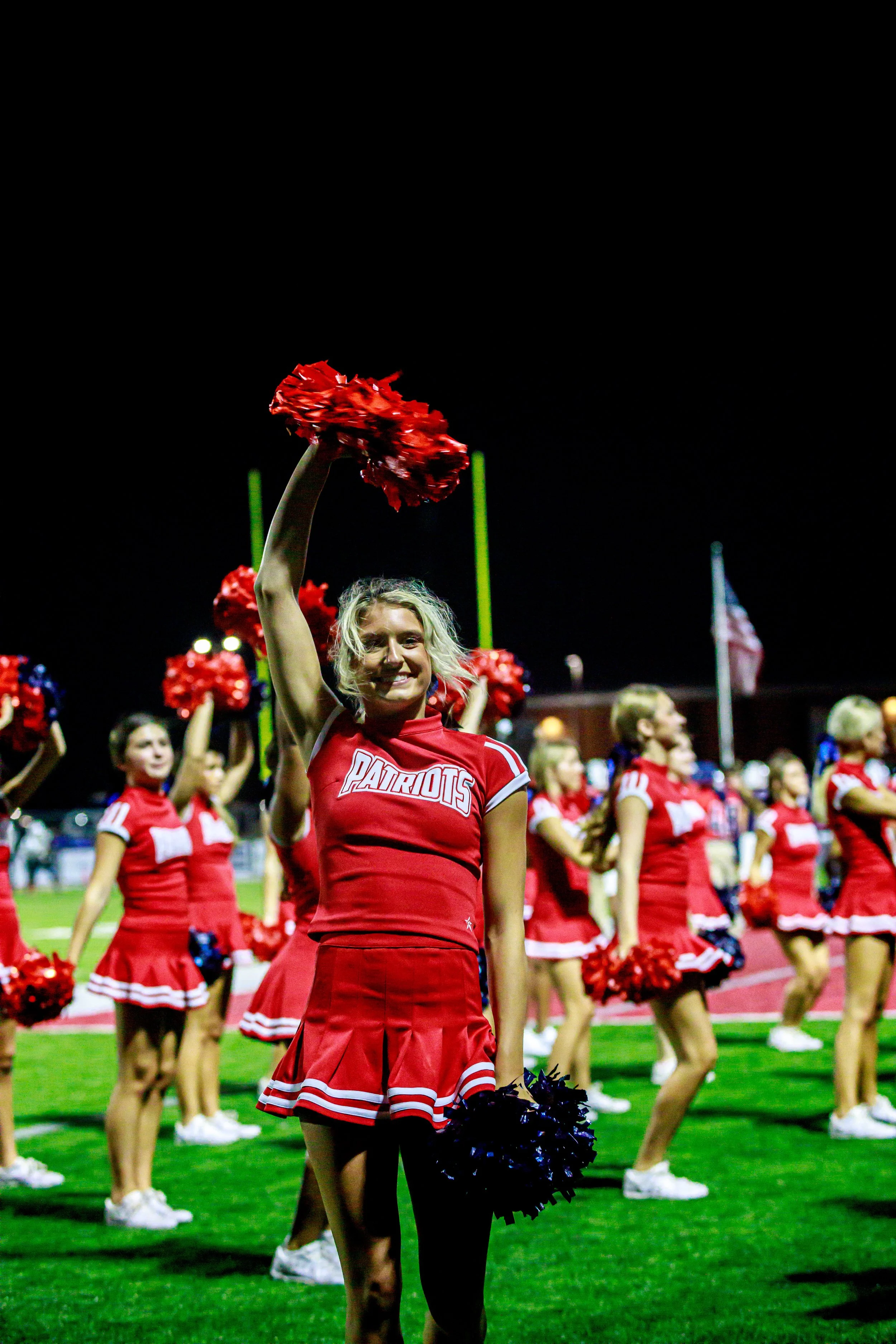 Cheerleader in a red Patriots uniform raises a red pom-pom during a nighttime football game, surrounded by teammates on the field.