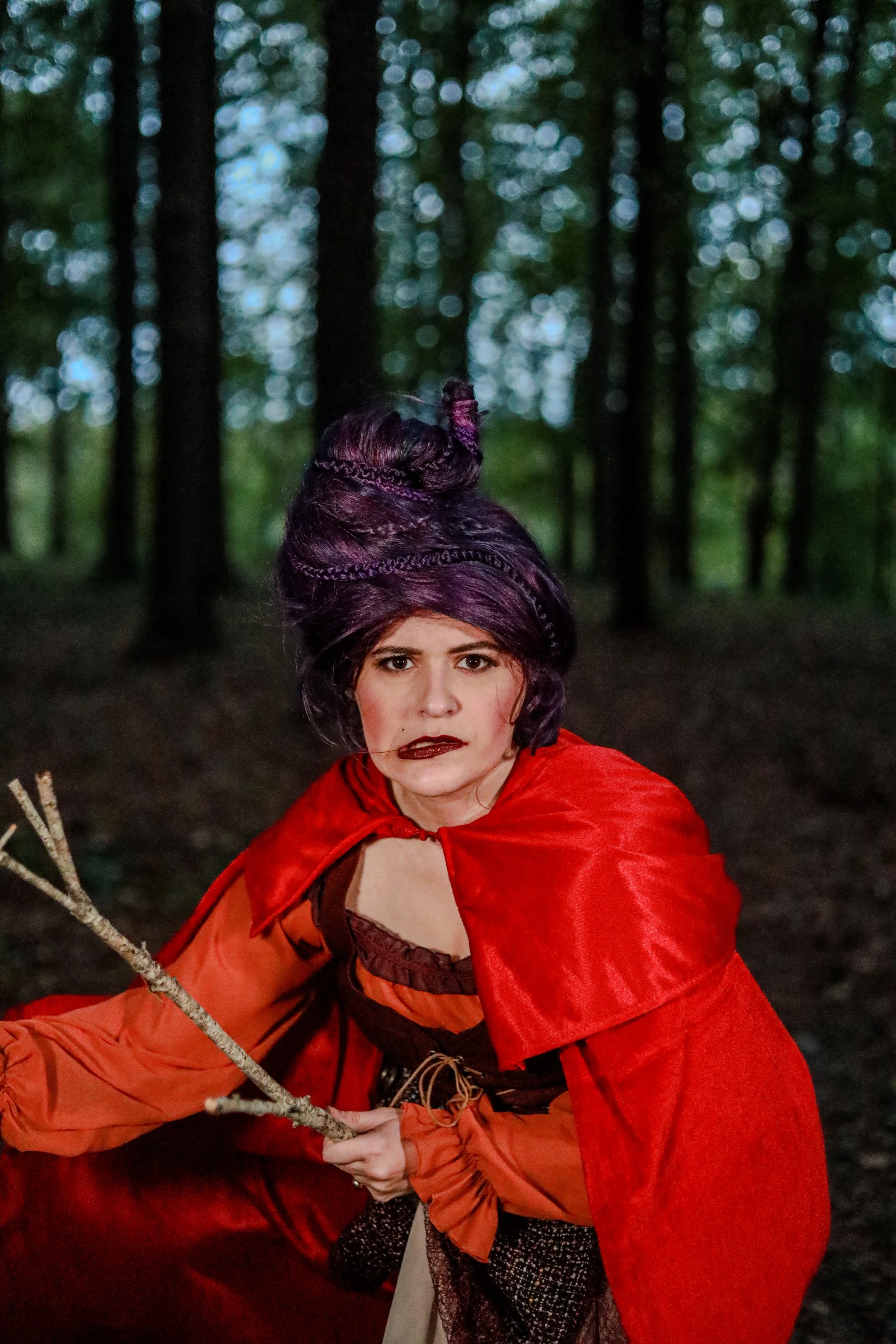 Woman in a red hooded cloak holding a branch in a dark, blurred forest