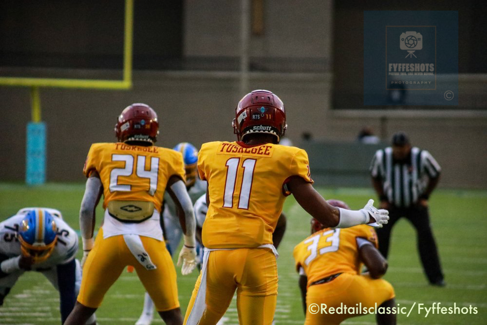 Tuskegee football players in gold uniforms and maroon helmets line up during a game.