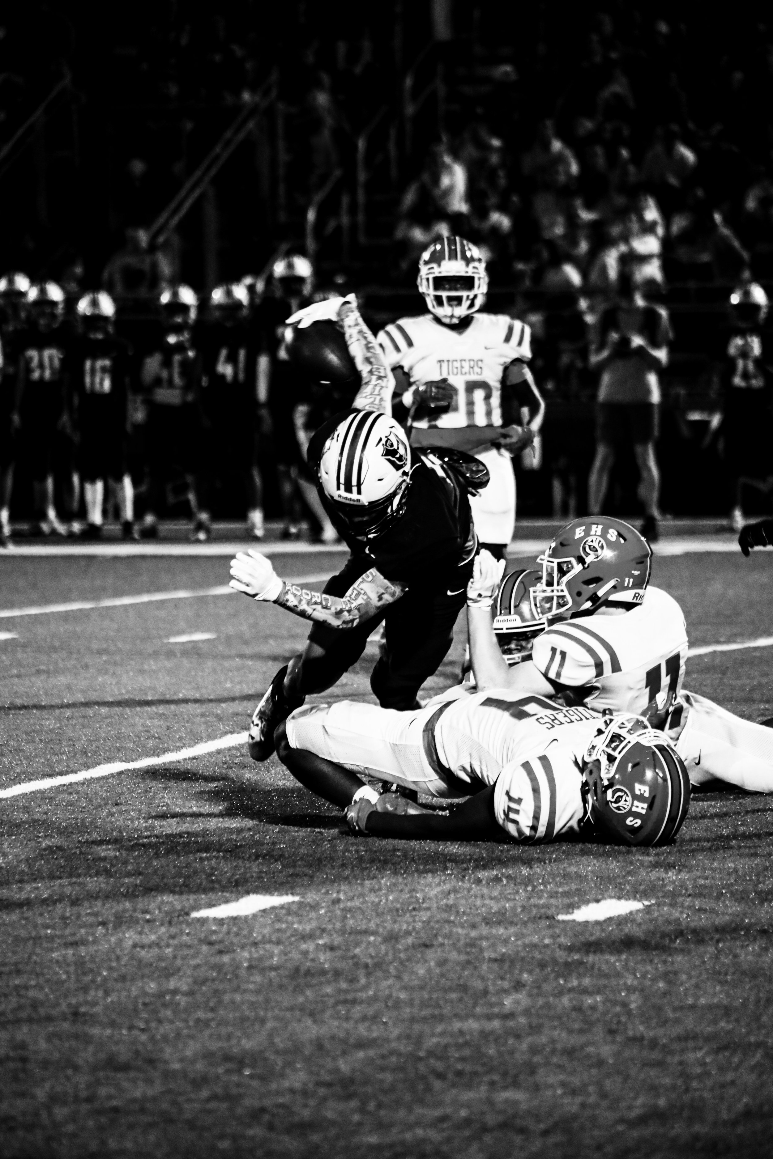 Black-and-white photo of high school football players tackling a Tigers ball carrier on the field during a nighttime game.