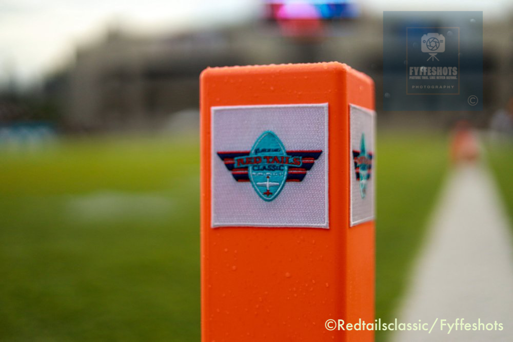 Close-up of an orange Red Tails Classic event sign beside a blurred grassy runway.