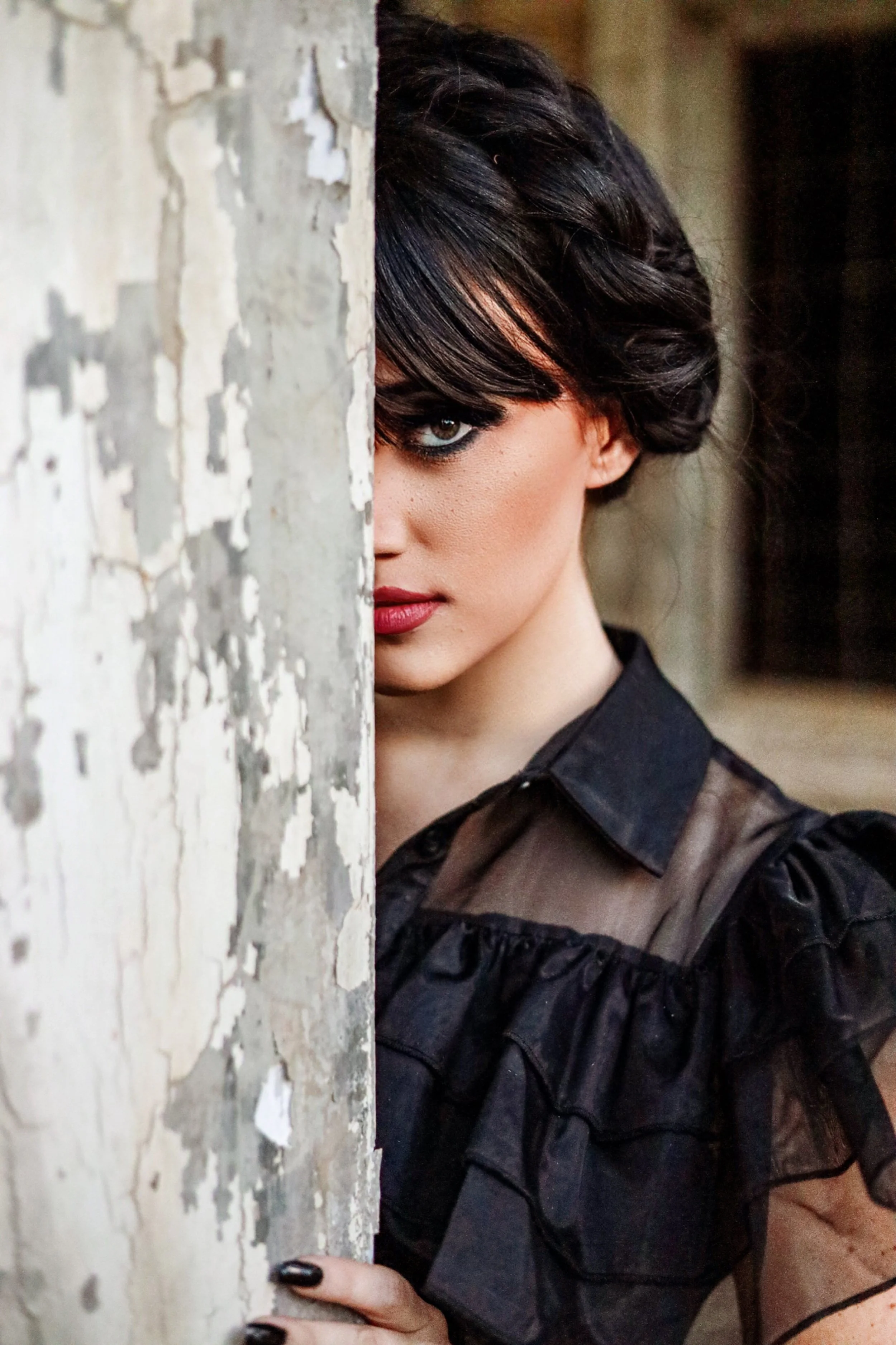 Close-up portrait of a woman with dark hair and red lipstick peering around a weathered wooden wall
