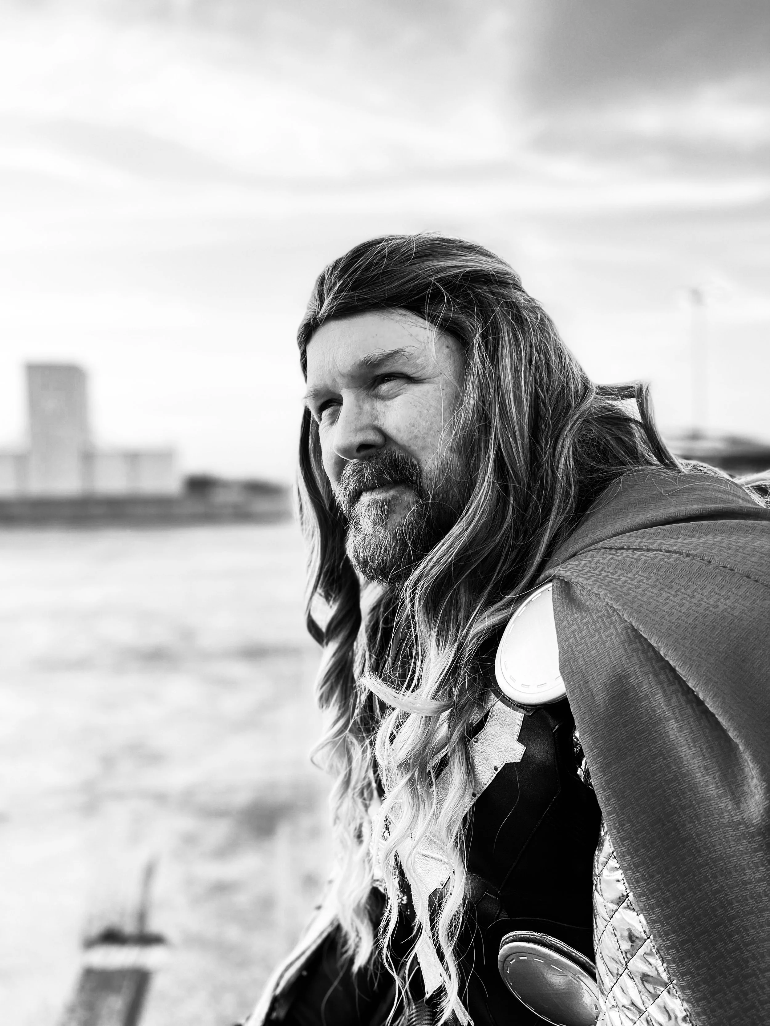Black-and-white close-up portrait of a long-haired, bearded man wearing a backpack outdoors.