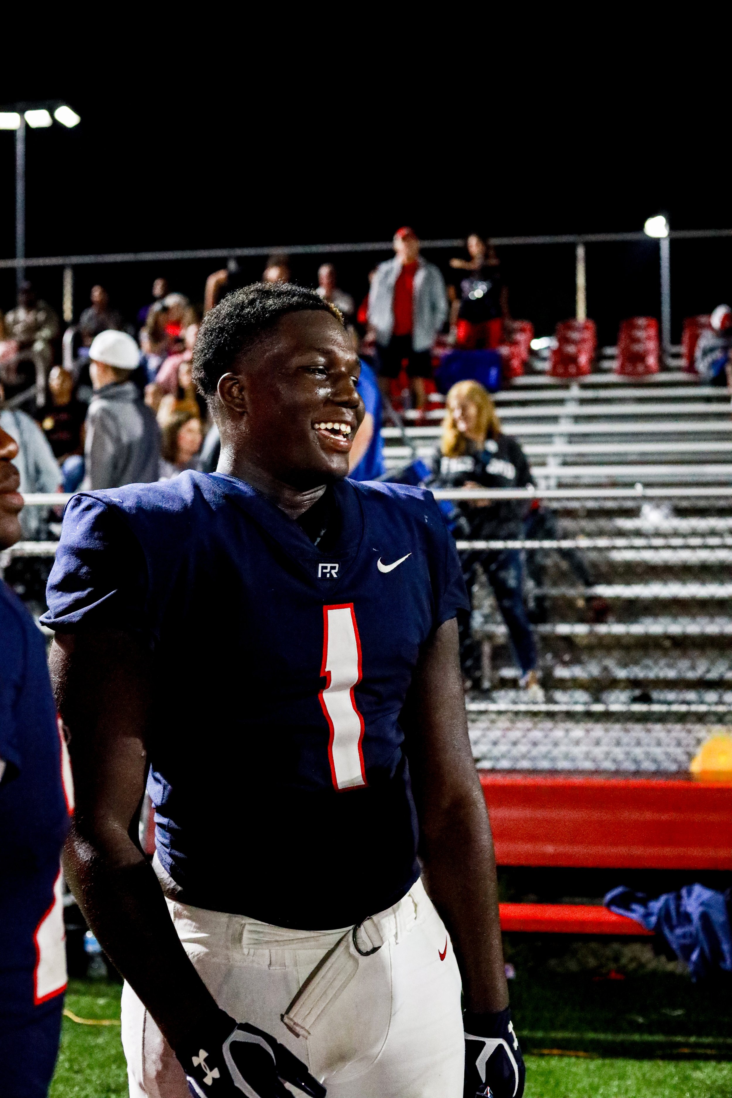 Smiling football player wearing a navy jersey with number 1 on a nighttime field, with spectators in the bleachers behind him