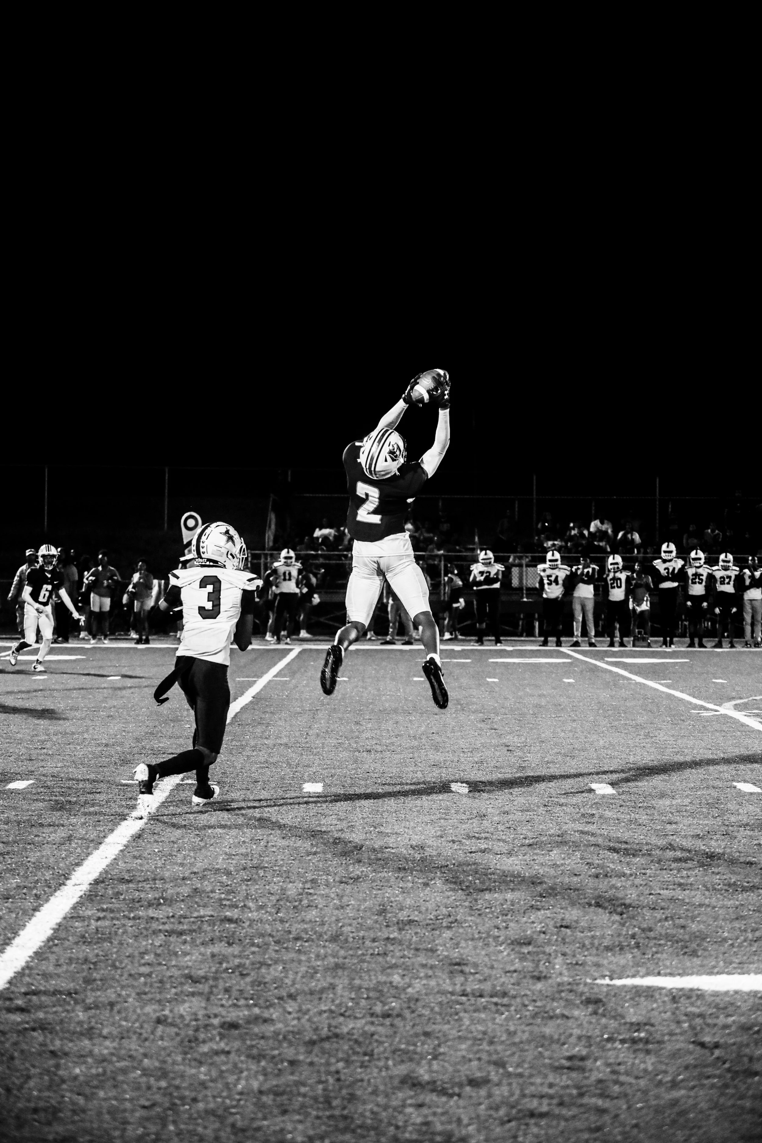 Black-and-white photo of a football player leaping to catch a pass as a defender pursues him on a high school field.