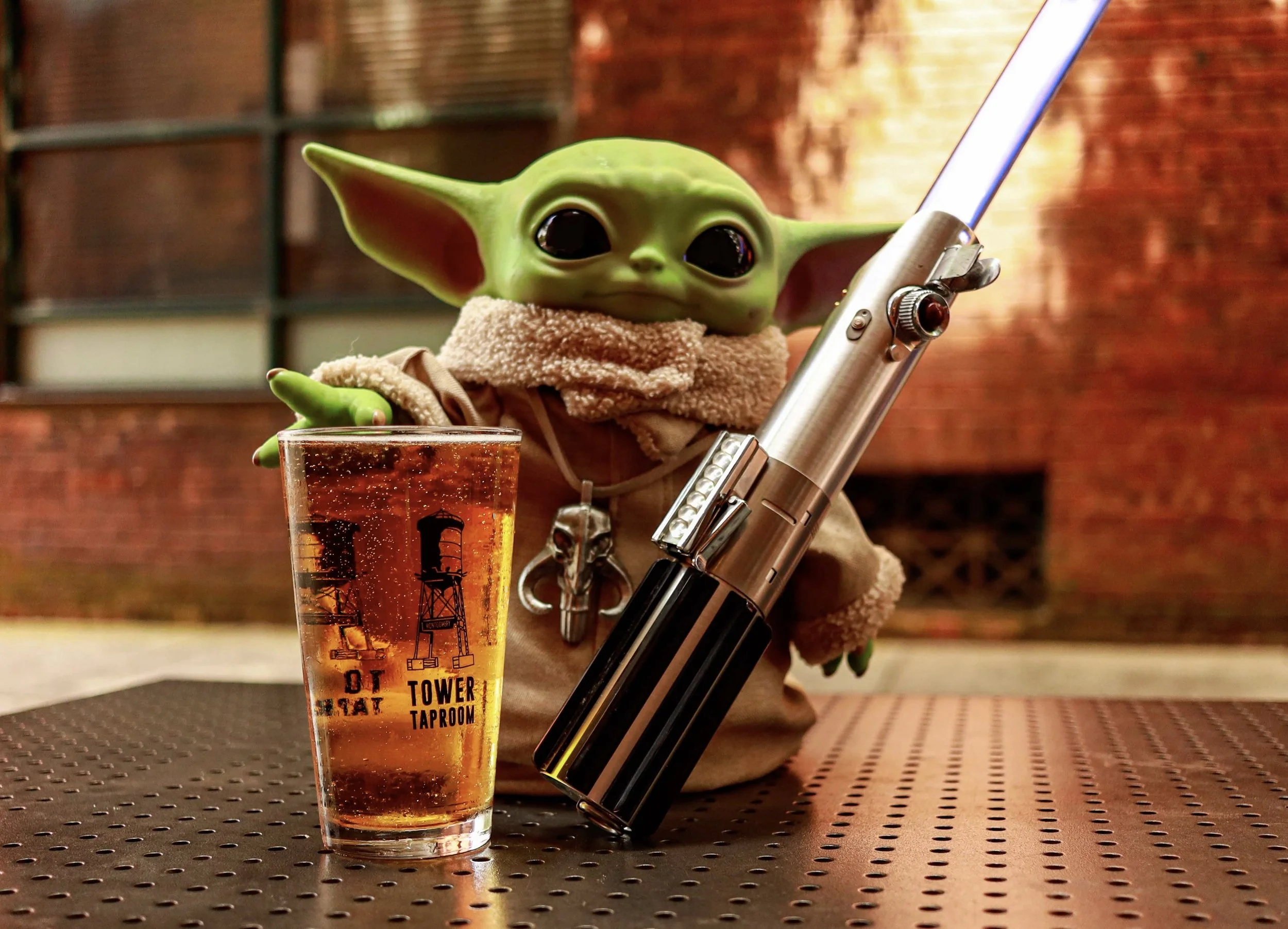Grogu figurine in a brown robe holding a lit lightsaber beside a glass of beer at a bar