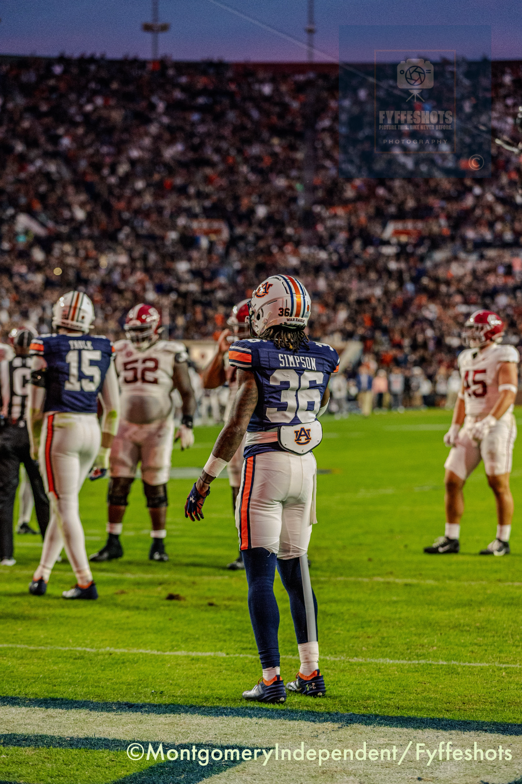 Auburn football player No. 35 stands on the field during a game against Alabama, with players and a blurred crowd in the background.