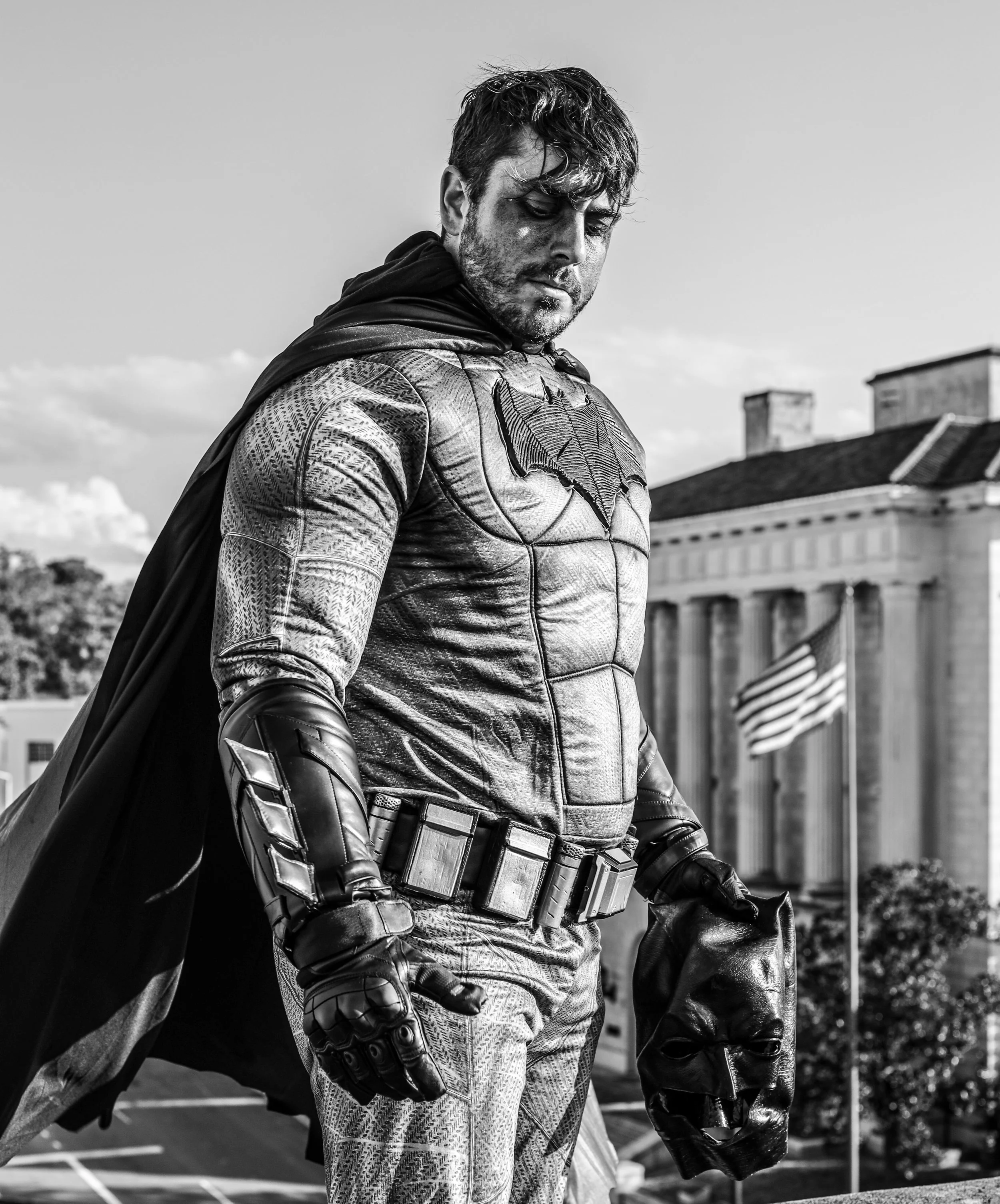 Black-and-white portrait of a Batman cosplayer in armored costume holding a Batman cowl on a rooftop