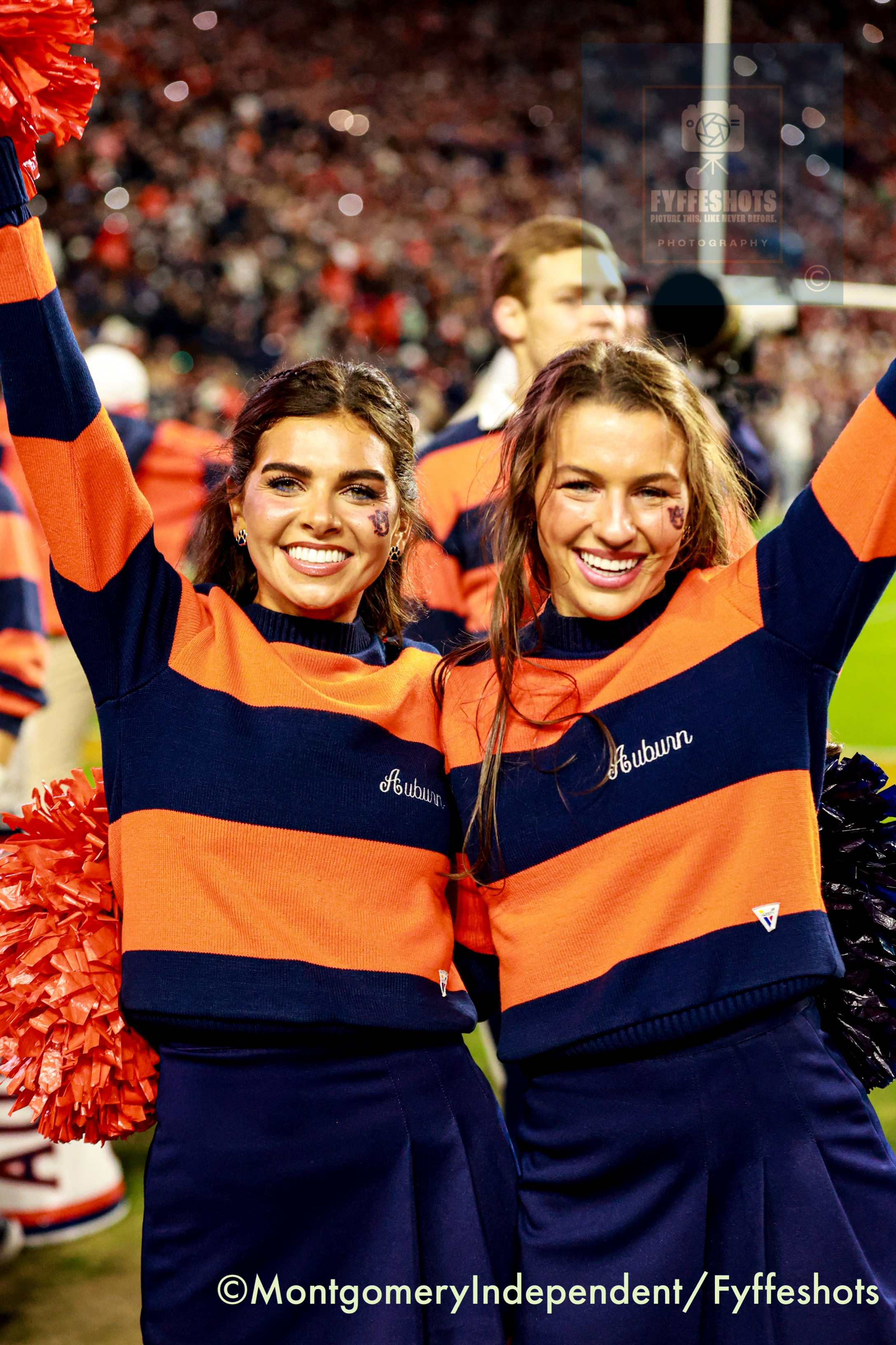 Two smiling Auburn cheerleaders in orange and navy uniforms pose with raised arms and pom-poms in a crowded stadium.