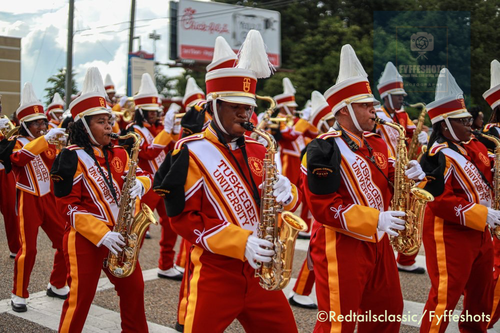 Tuskegee University marching band members in red-and-gold uniforms and white plumed hats playing saxophones in a parade