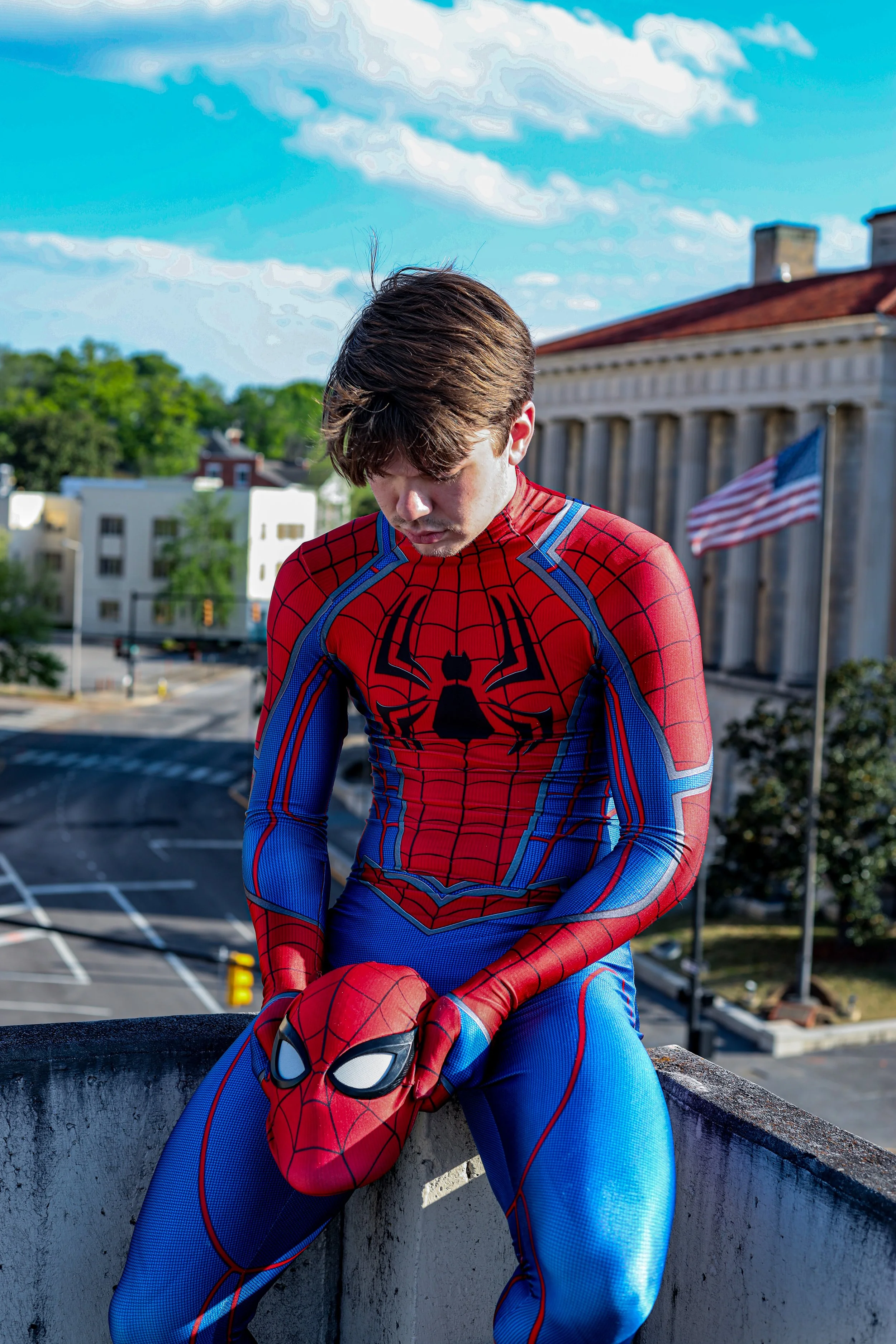 Person in a red and blue Spider-Man costume sitting on a rooftop in a city.
