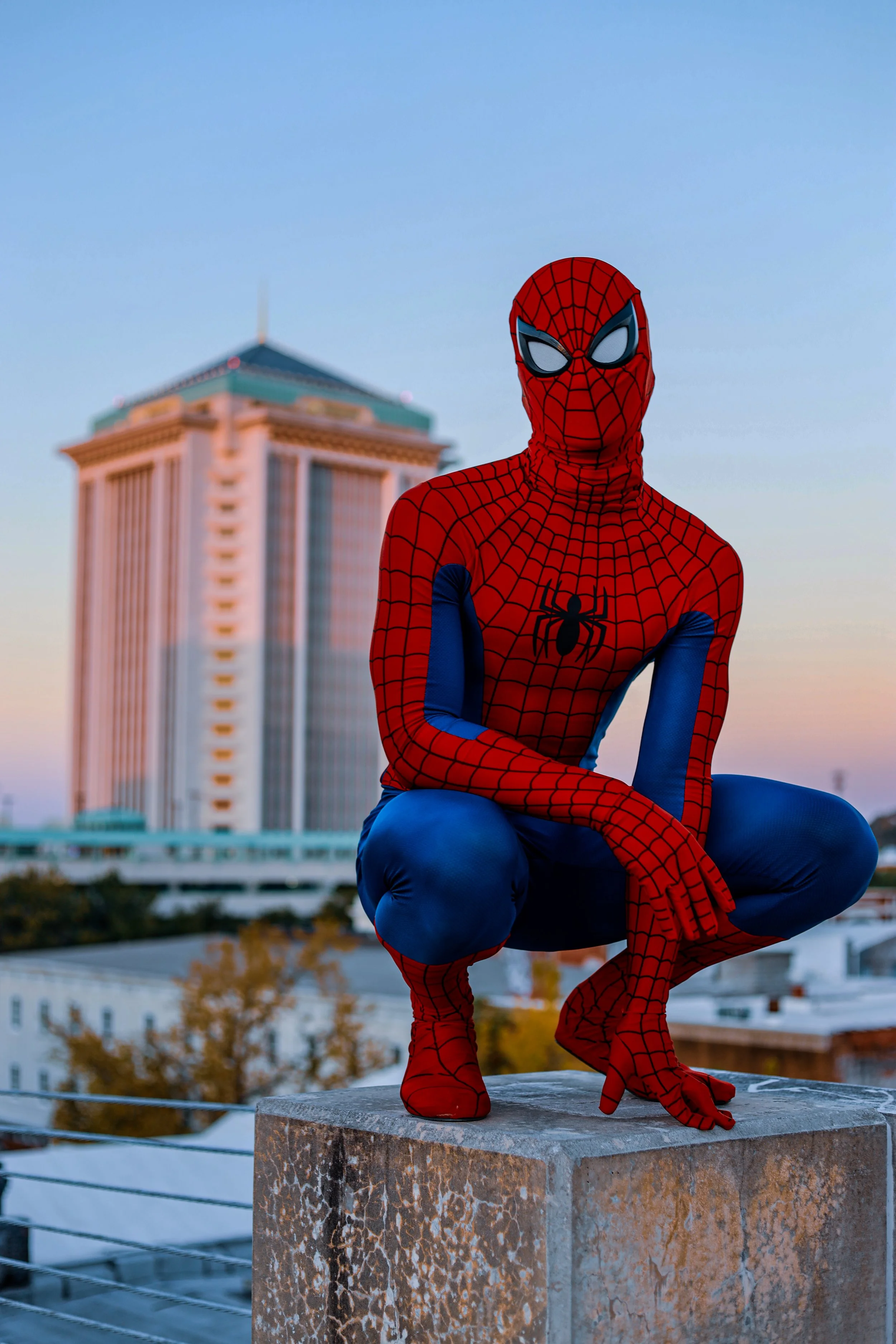 Person in a Spider-Man costume crouching on a rooftop with a city building in the background