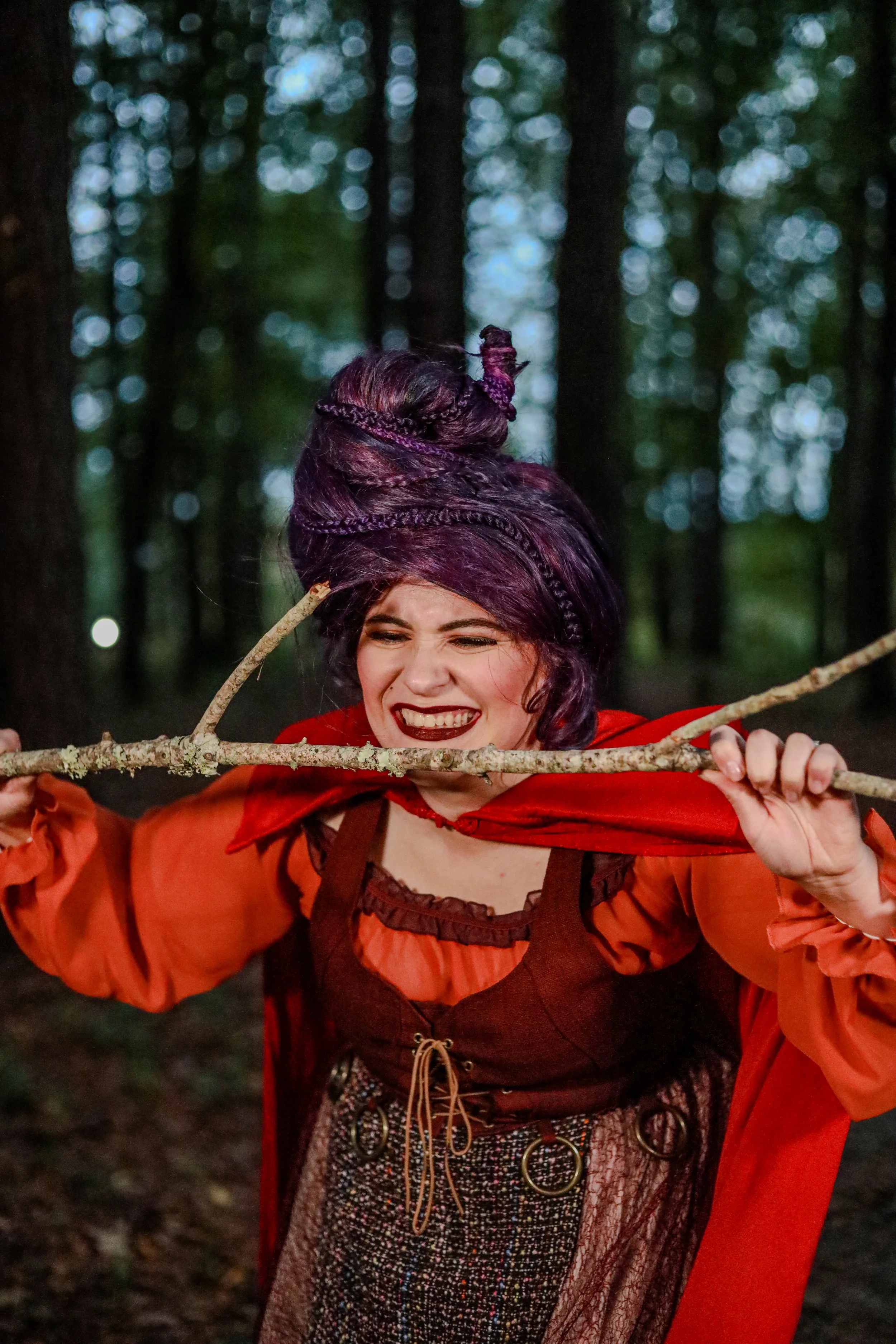 Smiling woman in a red hooded costume holding a wooden branch in a forest
