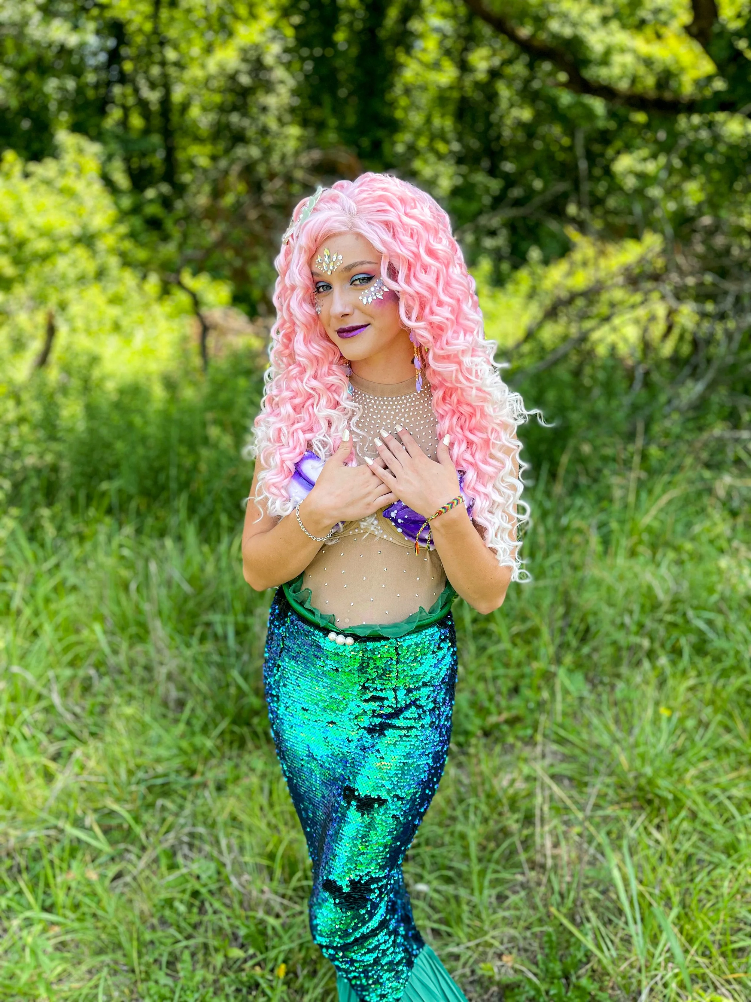 Person in a pink curly wig and jeweled makeup wearing a sparkling mermaid costume outdoors in a forest