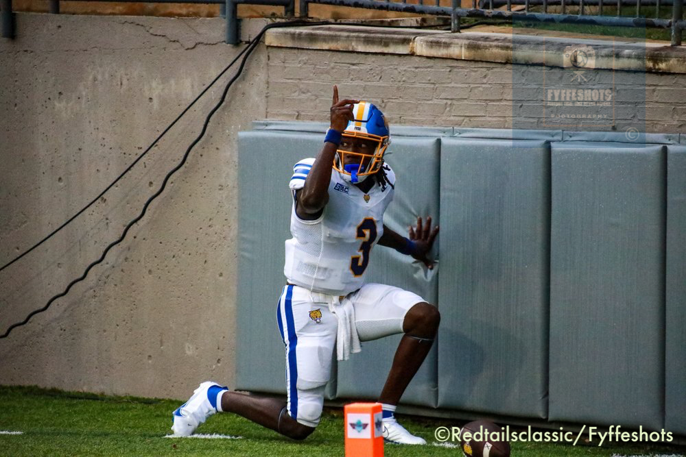College football player wearing a white and blue uniform kneels beside the end zone padding, raising one hand in celebration.