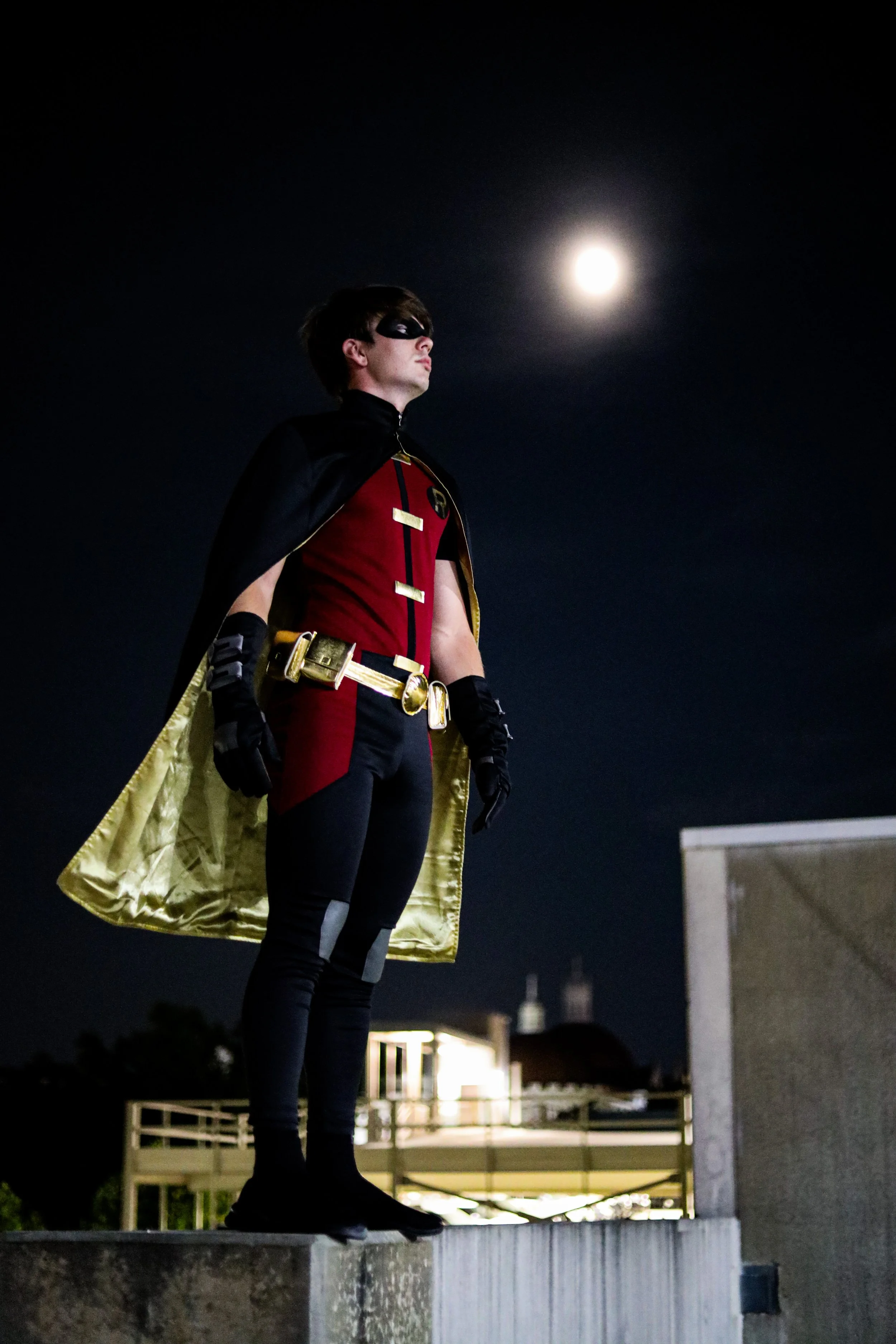 Masked person in a red-and-black superhero costume with a gold cape standing on a rooftop at night