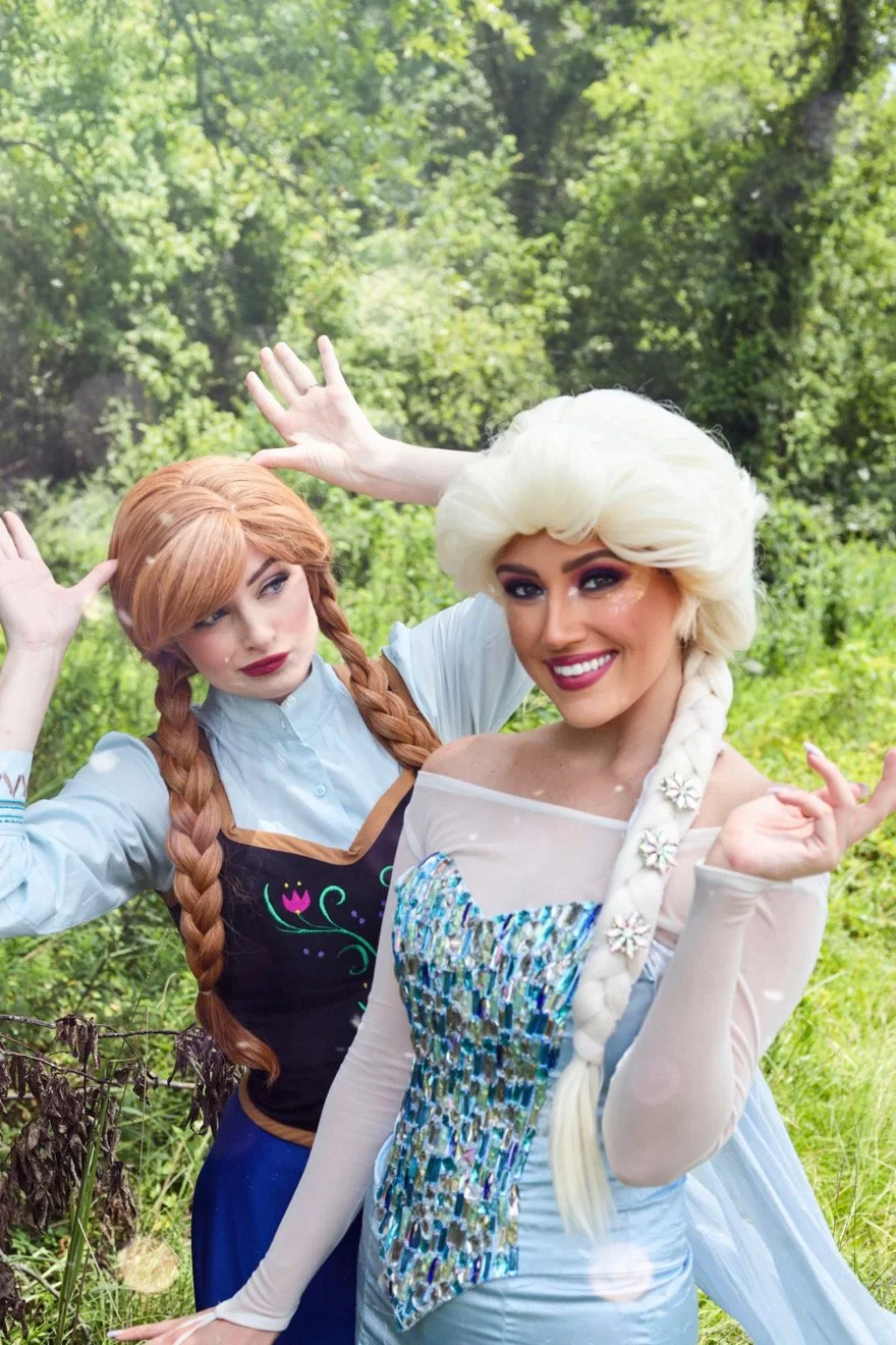 Two women in Elsa and Anna costumes posing outdoors in a forested park.
