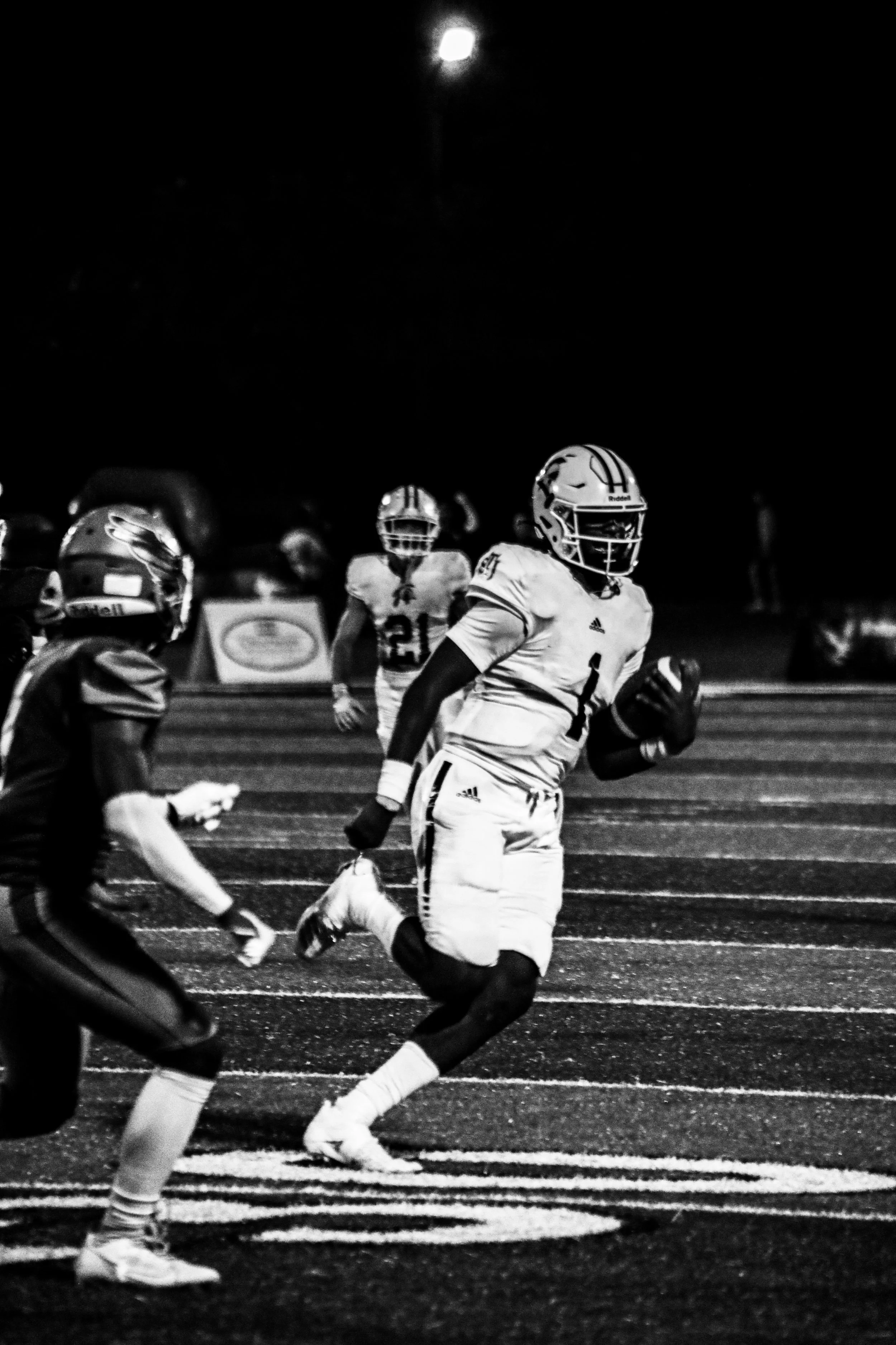 Black-and-white photo of a football player in a white uniform running with the ball as defenders pursue him under stadium lights.