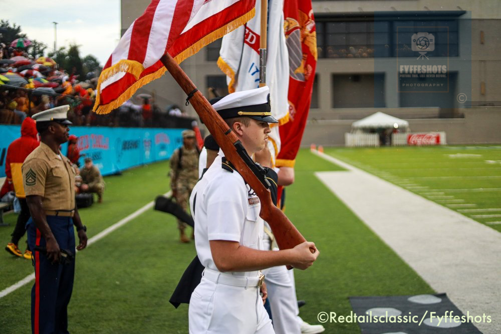 Military color guard officer carrying a rifle and American flag on a football field during a ceremony