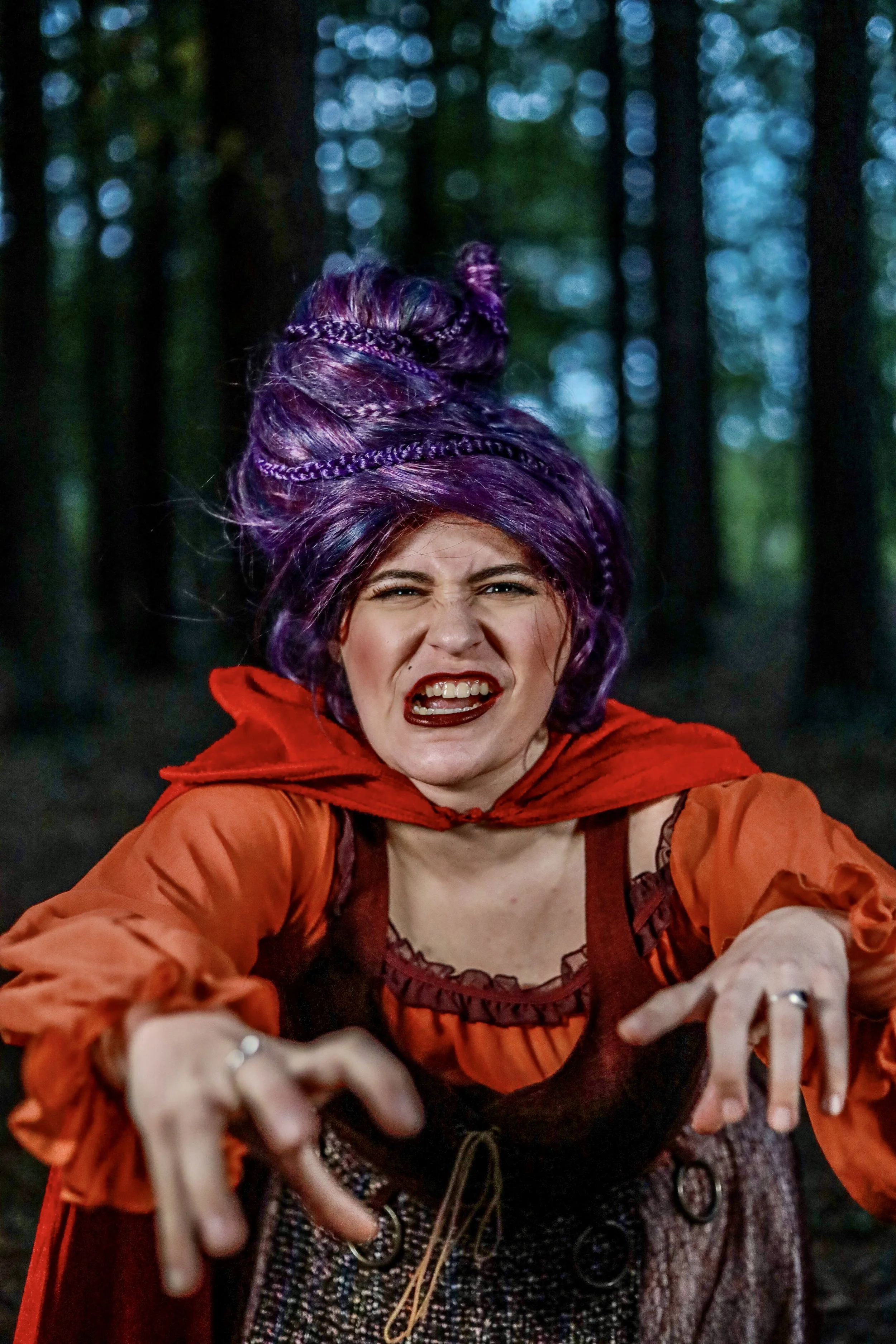 Smiling woman in a purple wig and red-and-orange costume posing in a dark forest