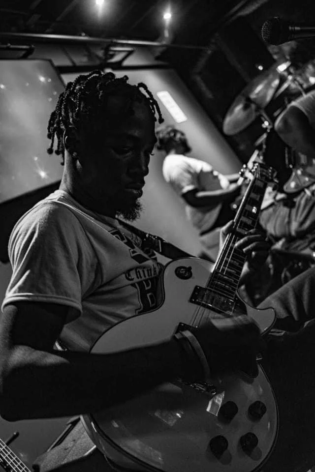 Black-and-white photo of a musician playing electric guitar on stage with a vocalist and bandmate in the background.