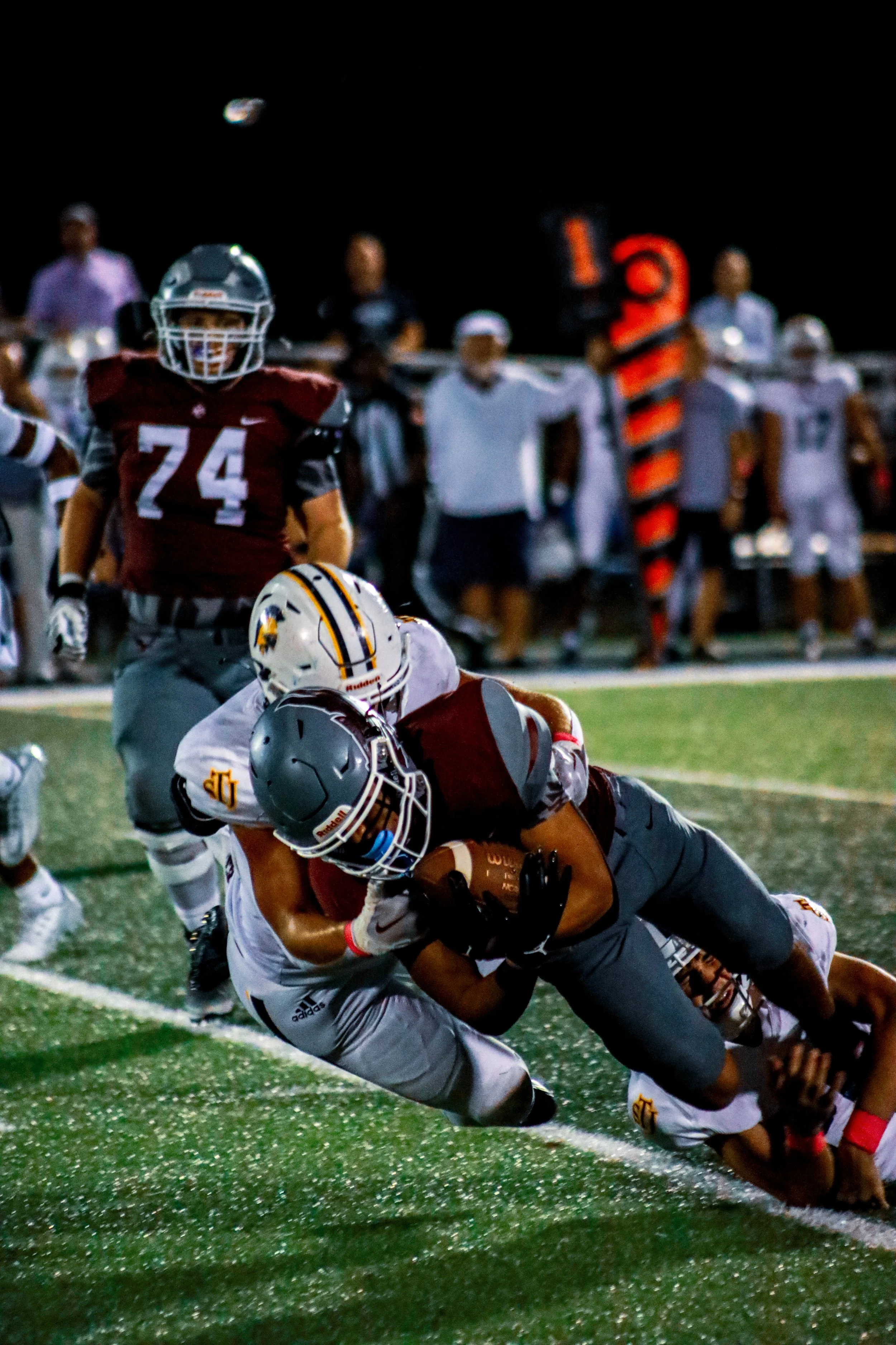 Football players tackle a maroon-clad ball carrier on a field during a nighttime game.