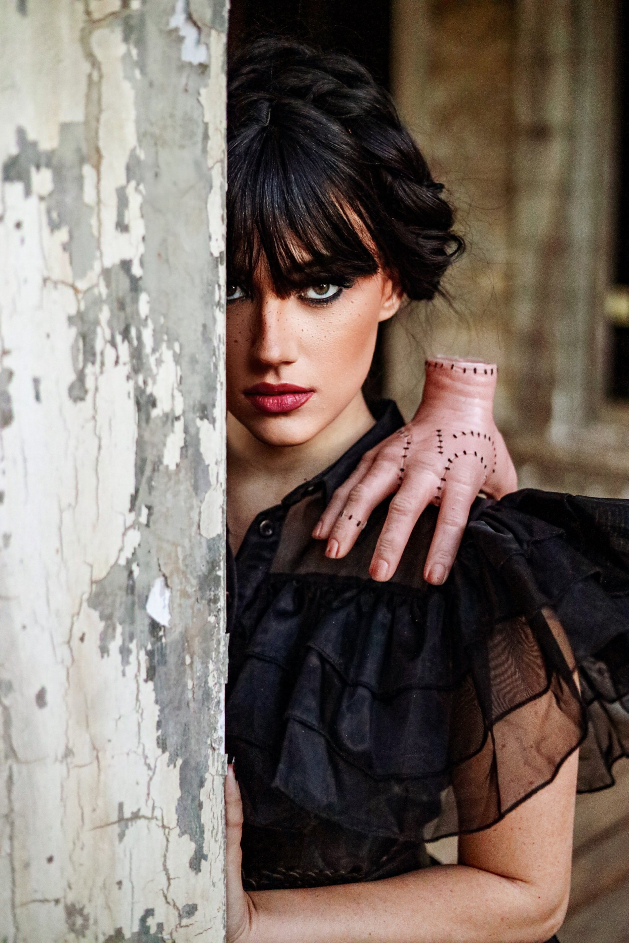 Gothic woman with black hair, dramatic eye makeup, and red lipstick peeking around a weathered wall