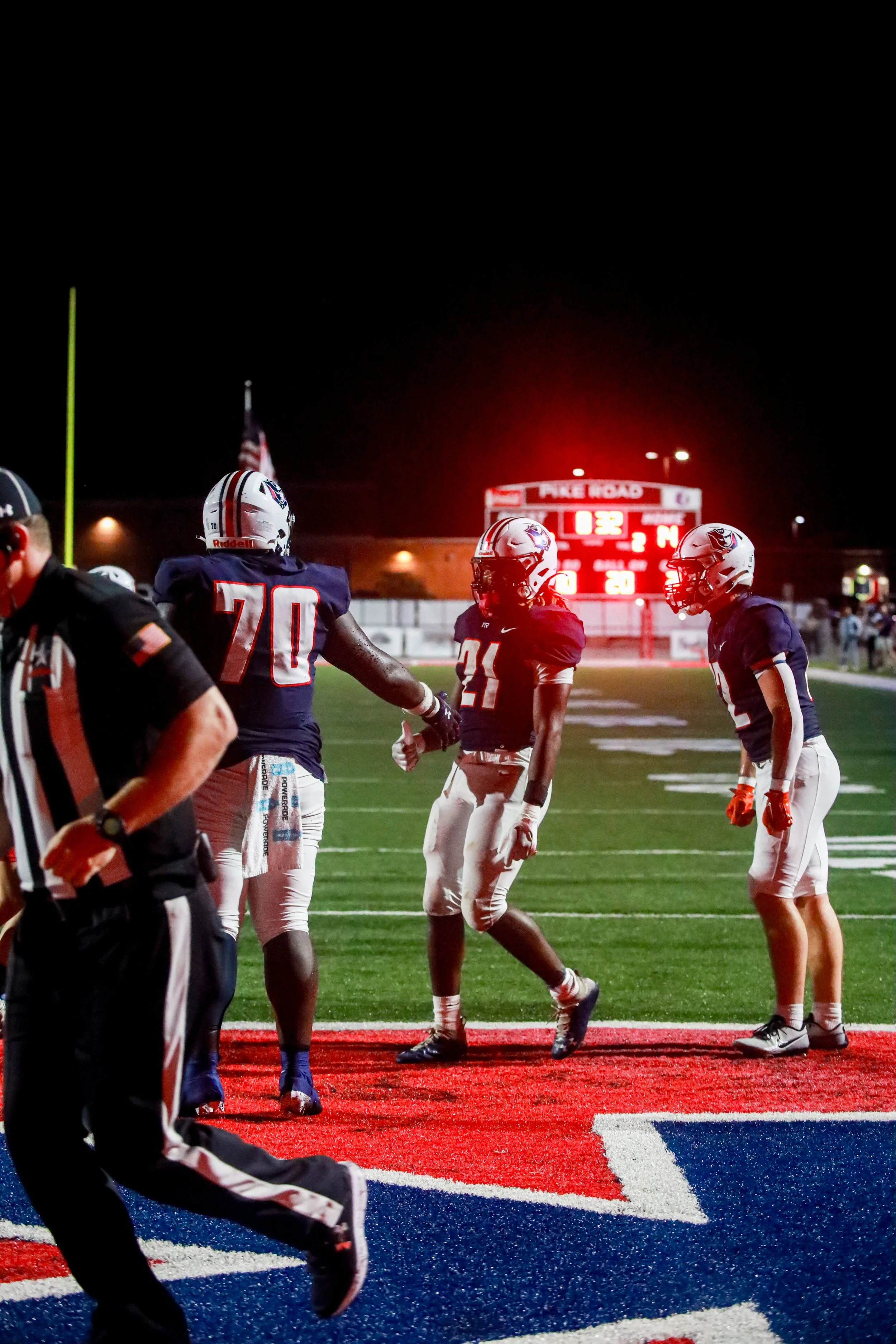 High school football players in navy uniforms and white helmets stand on a red-and-blue end zone at night, with a glowing scoreboard behind them.