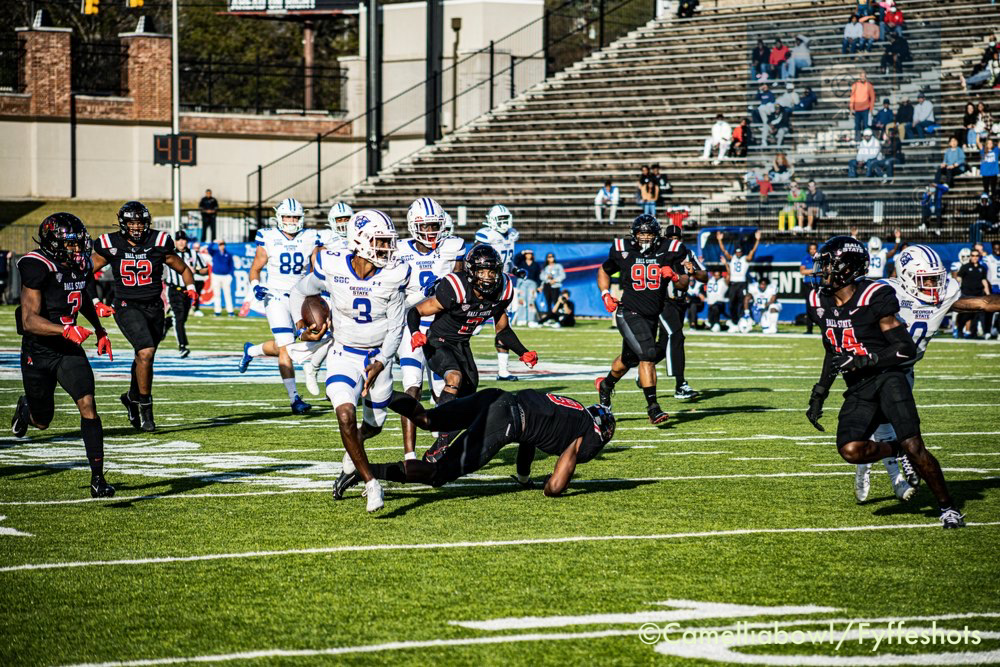 College football player in white jersey runs with the ball as opposing black-uniformed defenders pursue on a field.