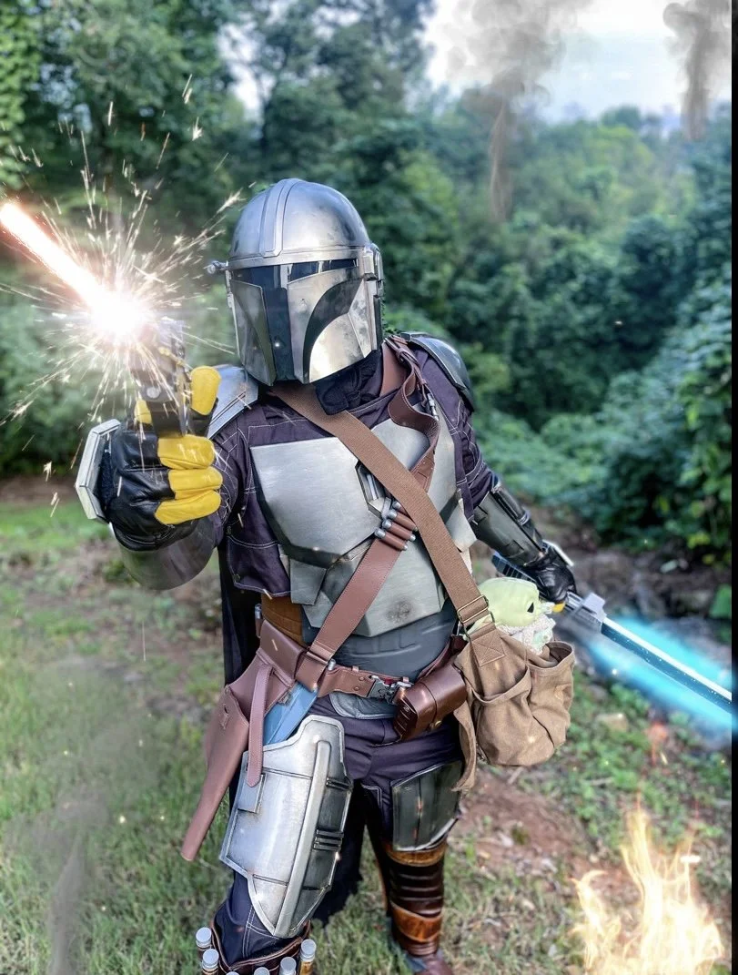 Cosplayer in Mandalorian armor holding a glowing blue lightsaber and yellow energy device in a forest