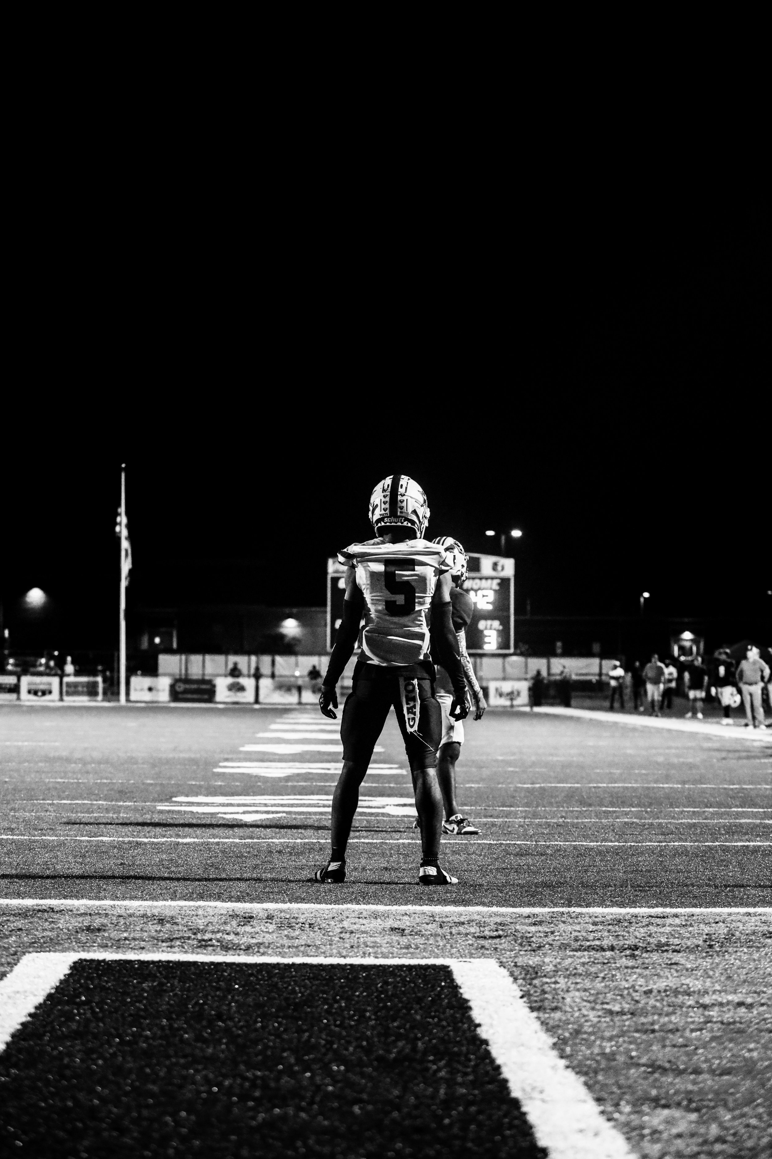 Black-and-white photo of two football players standing on a field at night, with a scoreboard and spectators in the background.