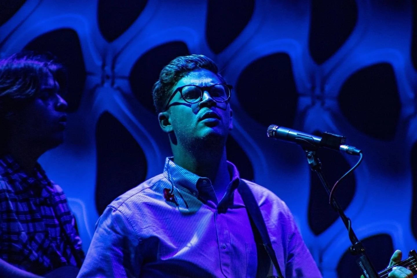 Musician wearing glasses sings into a microphone under blue stage lighting during a live performance.