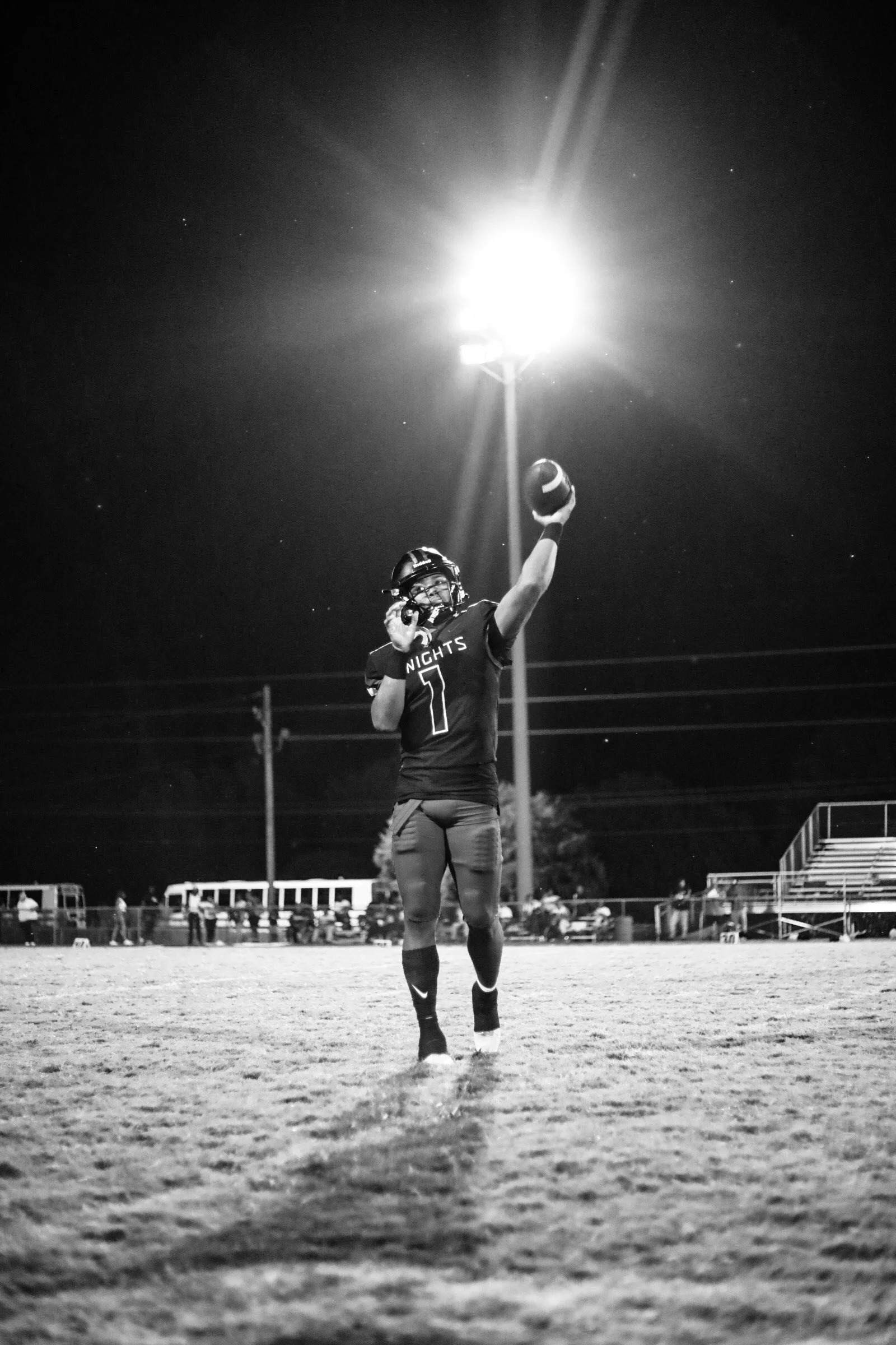 Black-and-white photo of a football player wearing jersey number 1 and raising a football under stadium lights at night