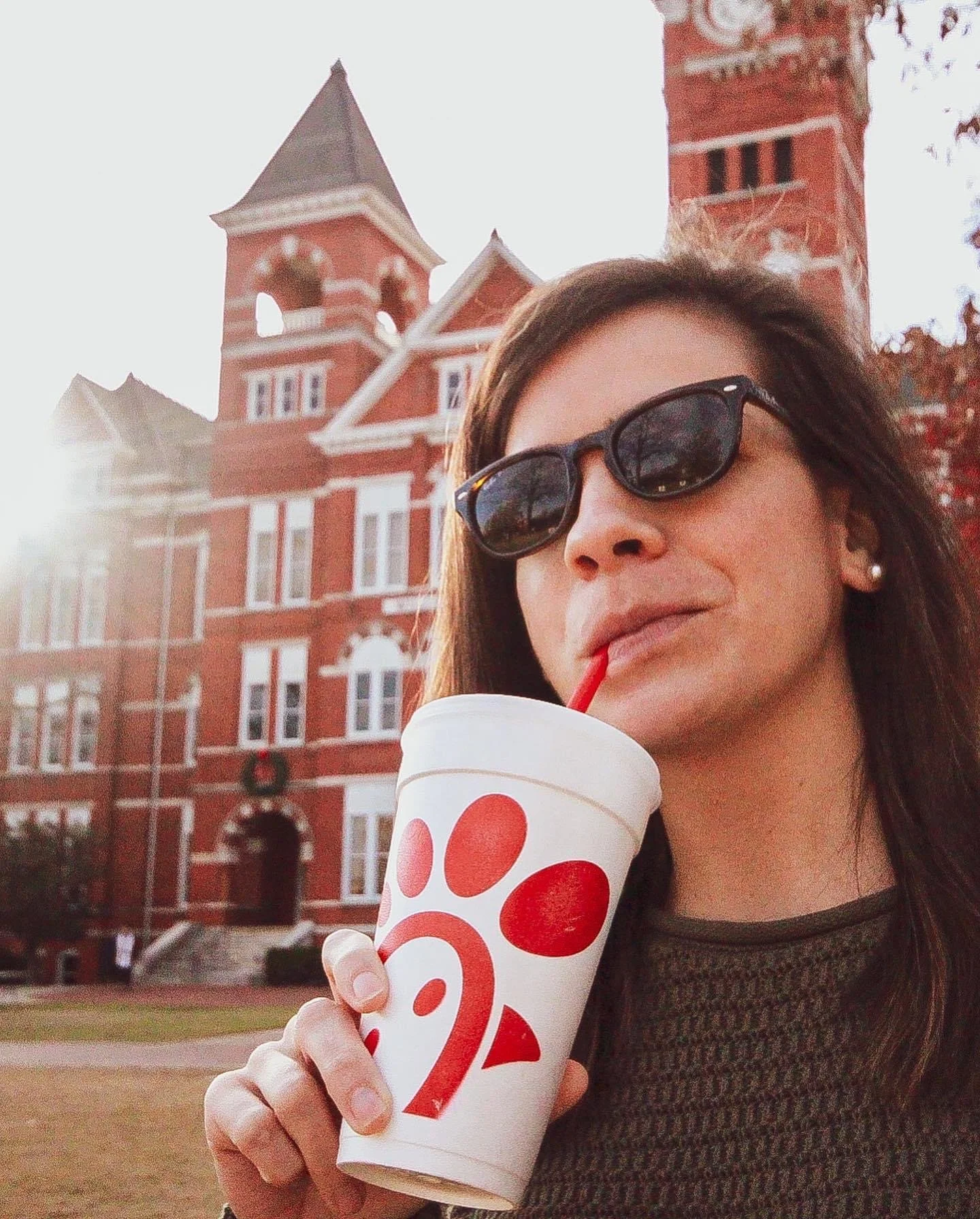 Woman wearing sunglasses holds a Chick-fil-A drink in front of a historic red-brick building.