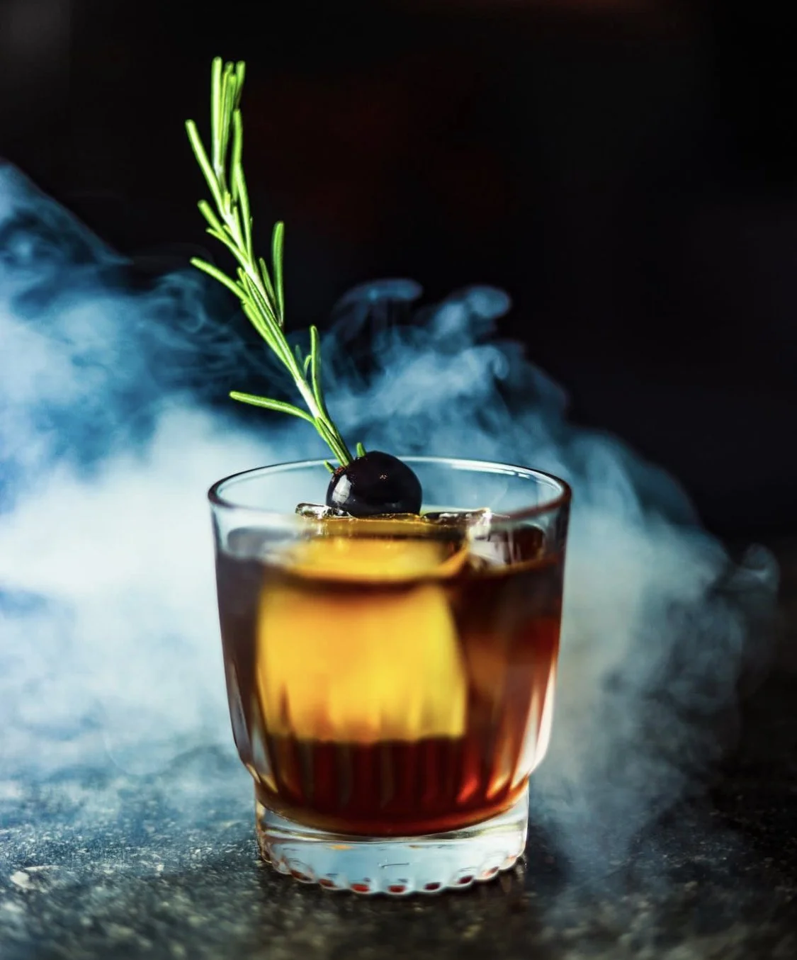 Dark amber cocktail in a rocks glass garnished with a cherry and rosemary sprig, surrounded by blue smoke.