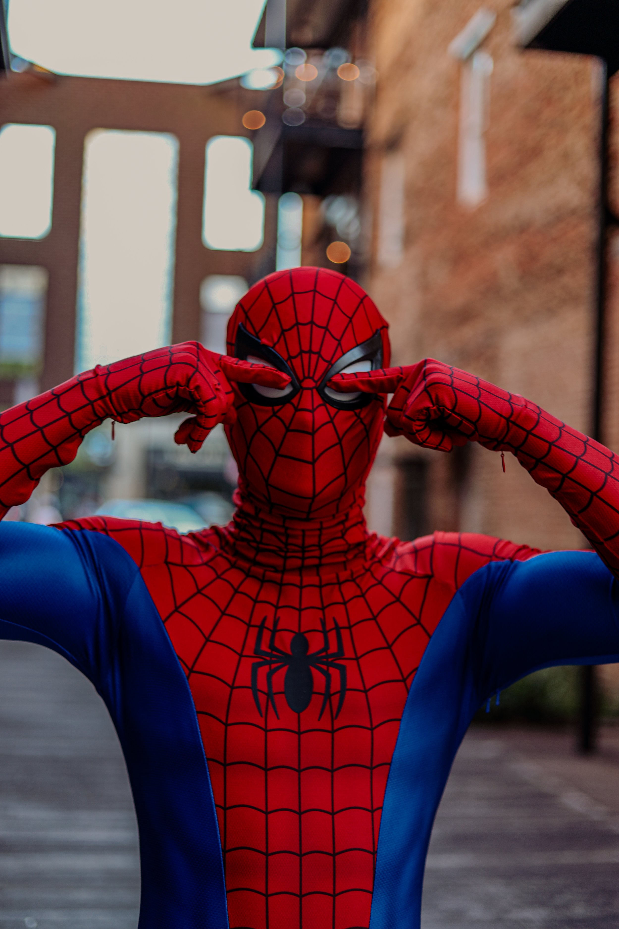 Person in a red-and-blue Spider-Man costume posing with hands raised in front of a blurred city street.