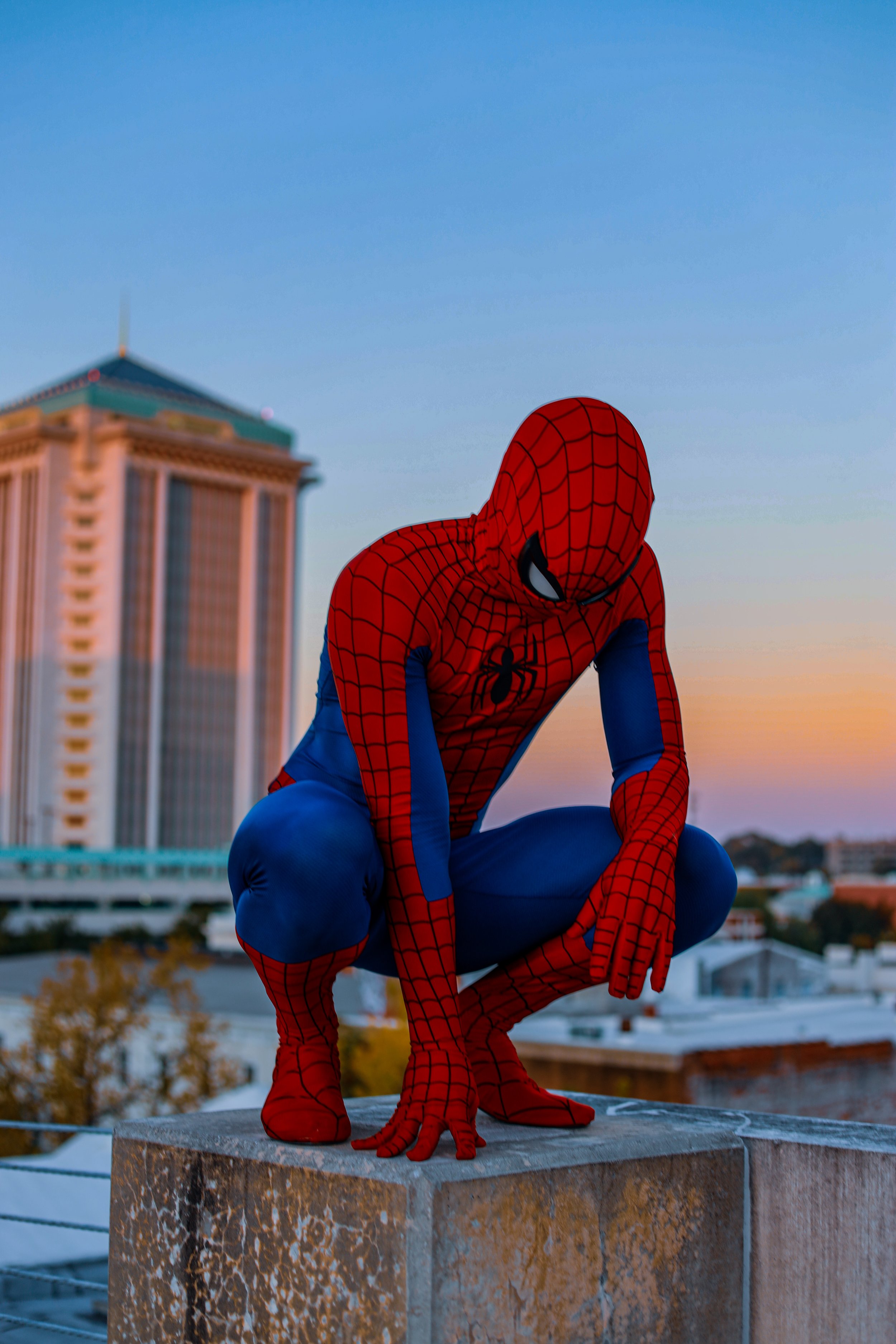 Person in a red-and-blue Spider-Man costume crouching on a rooftop with a city skyline behind them
