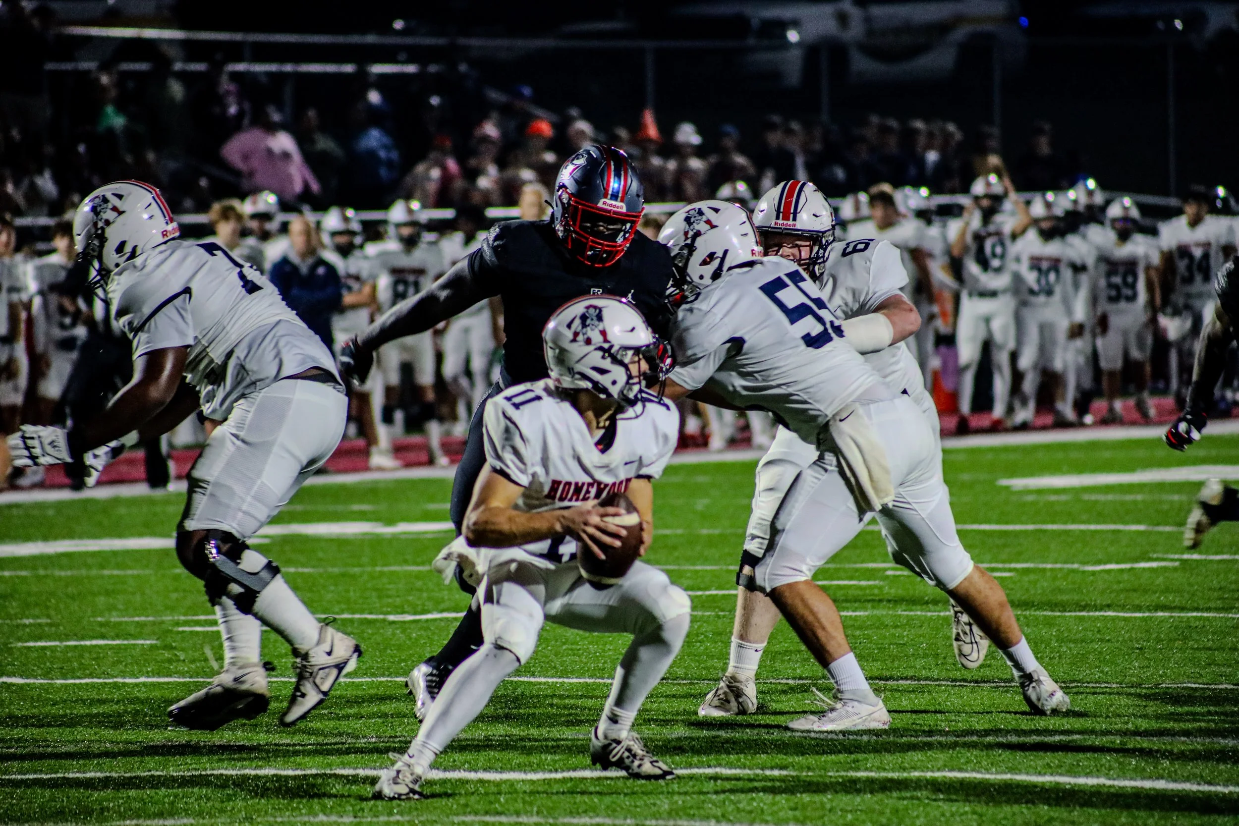 High school football quarterback runs with the ball as teammates block a defender under stadium lights