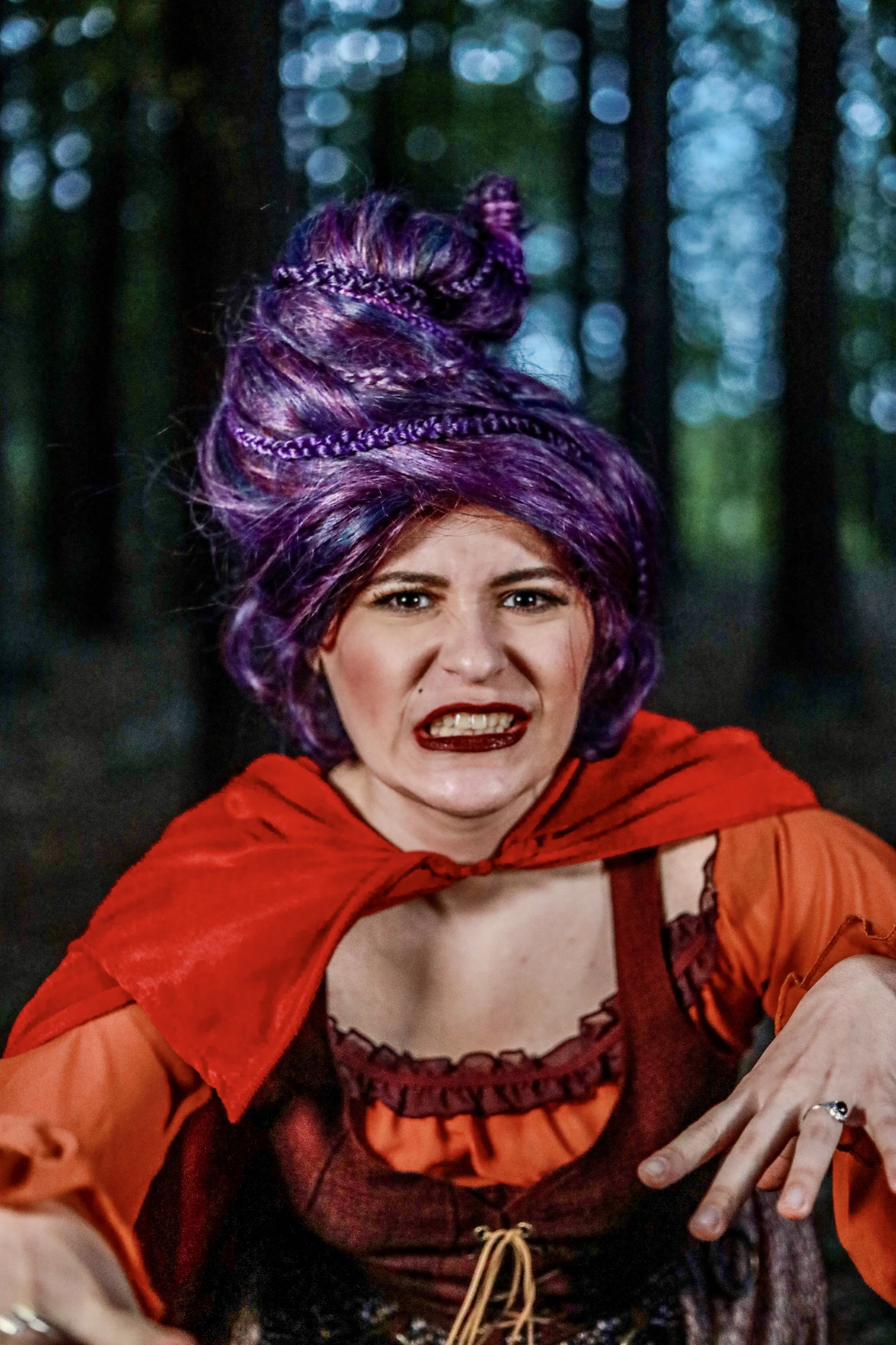 Smiling woman in a purple witch hat and red-orange costume against a dark, blurred background